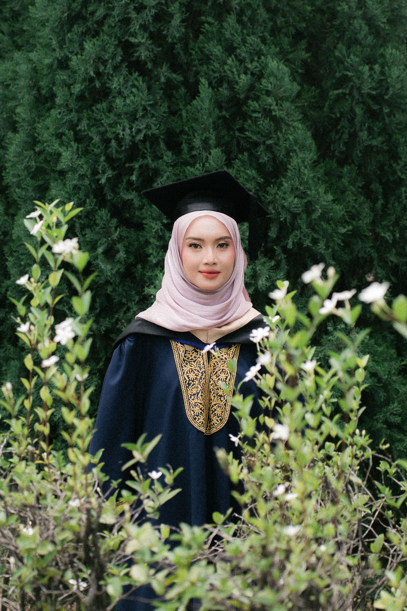 A young woman in graduation gown and cap wearing a pink hijab stands among green trees and white flowers.