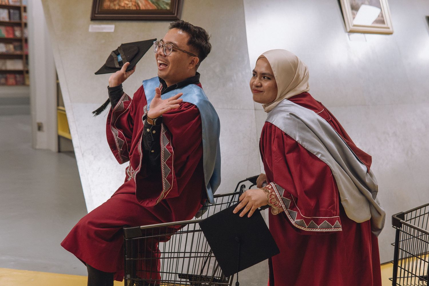 Two graduates in maroon academic regalia pose playfully with a shopping cart in a hallway.