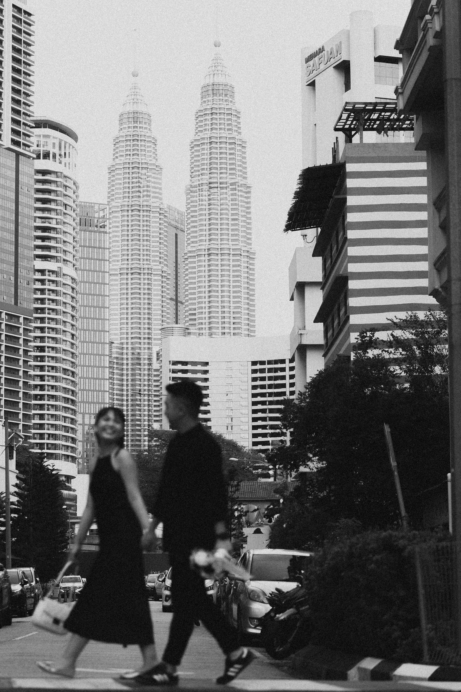 A black and white cityscape featuring the iconic Petronas Towers in Kuala Lumpur with pedestrians walking in the foreground.