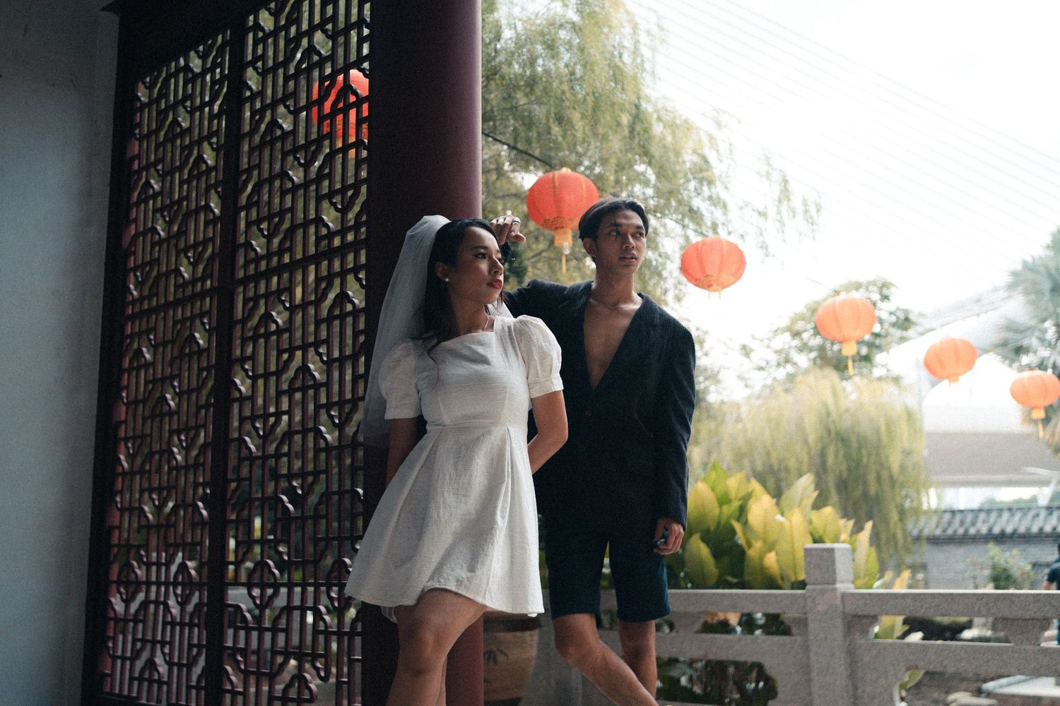 Couple in wedding attire posing near decorative wall with orange lanterns.