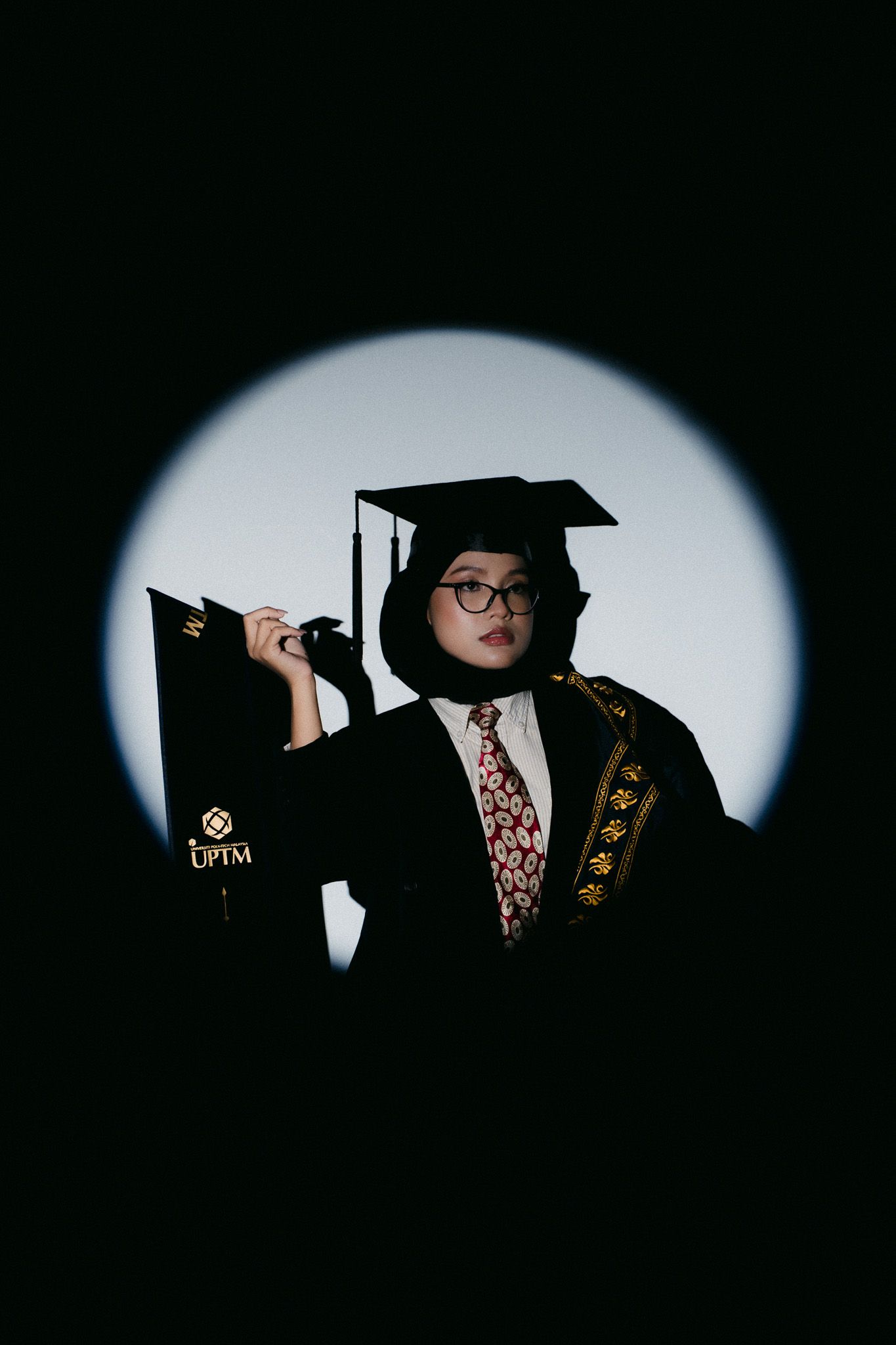 Graduate in black cap and gown with hijab holds diploma under spotlight.