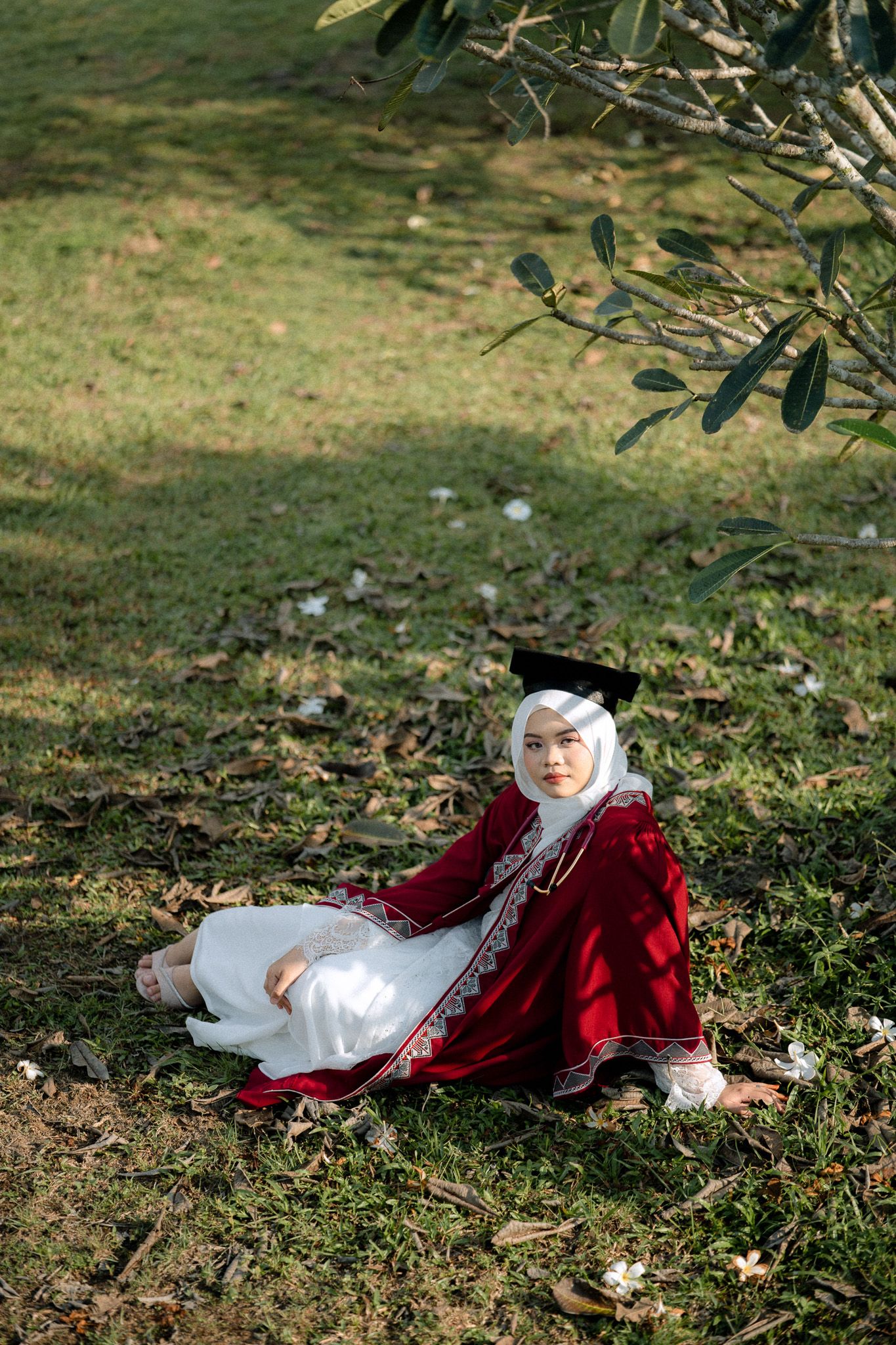 Muslim woman in red and white dress with hijab and graduation cap sitting on grass under a tree outdoors.