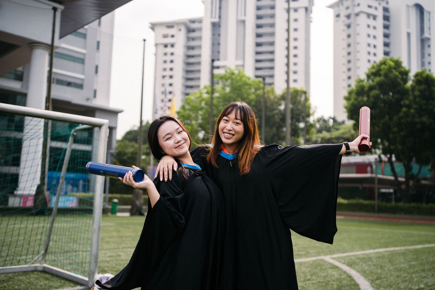 Graduates in black academic robes celebrating together on a sports field with high-rise buildings in background.