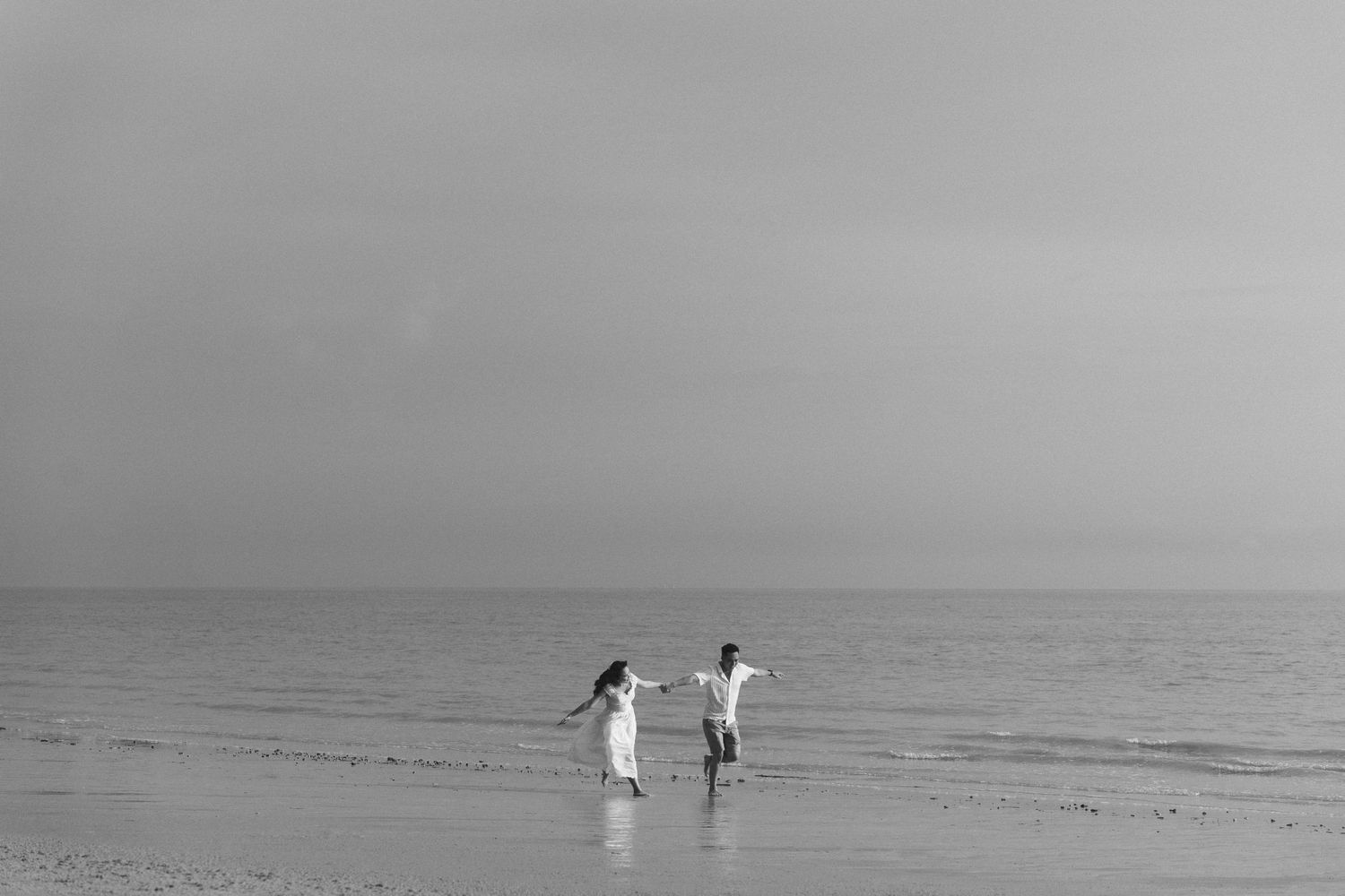 Black and white photo of two people running joyfully along beach shoreline.