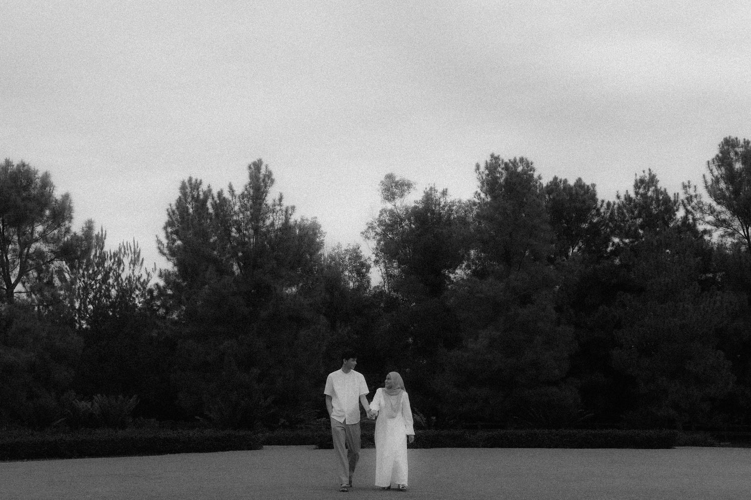 Two people in white outfits stand together against a backdrop of trees in a black and white photo.