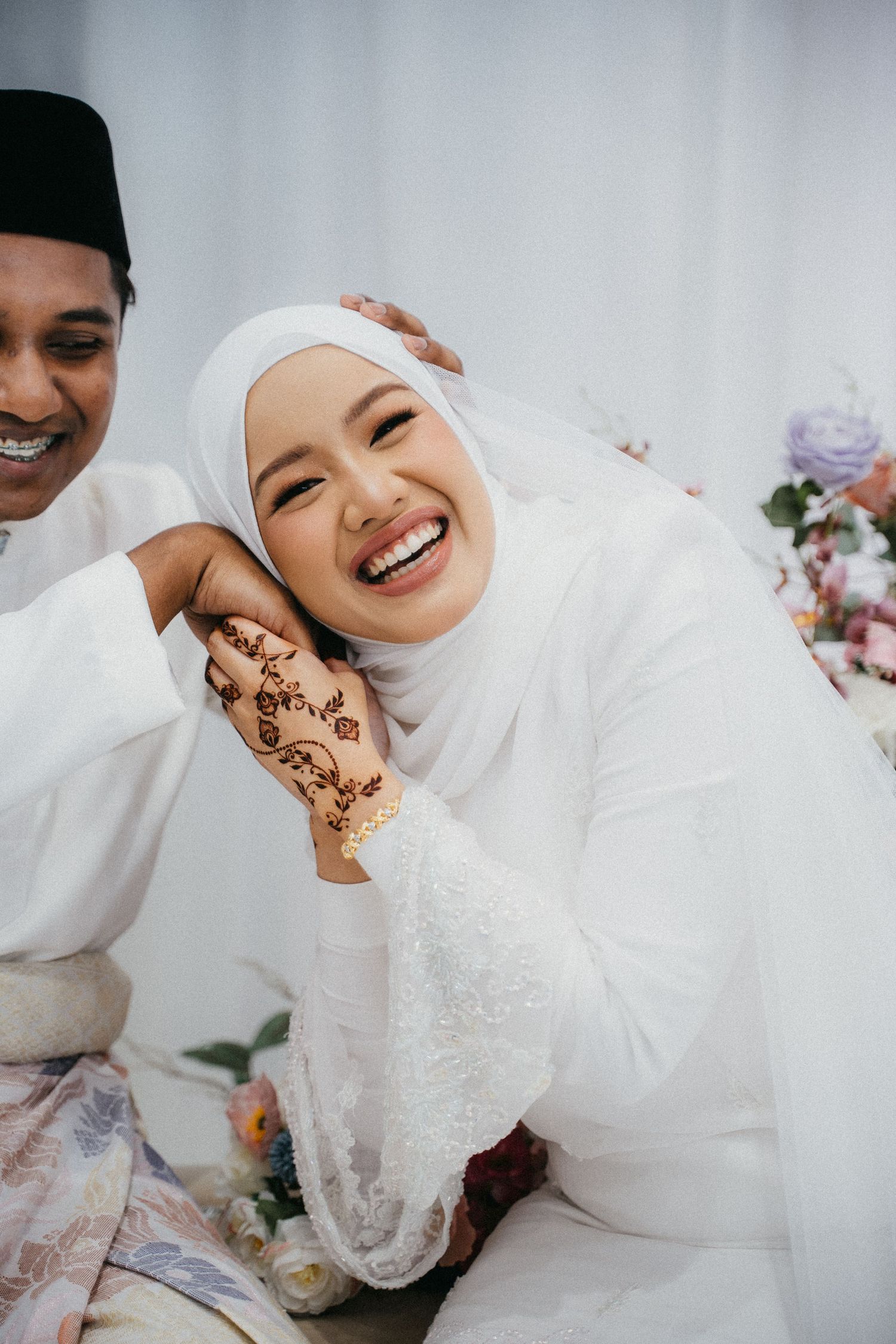 A joyful couple in white traditional wedding attire share loving moments with elegant henna decorations visible.