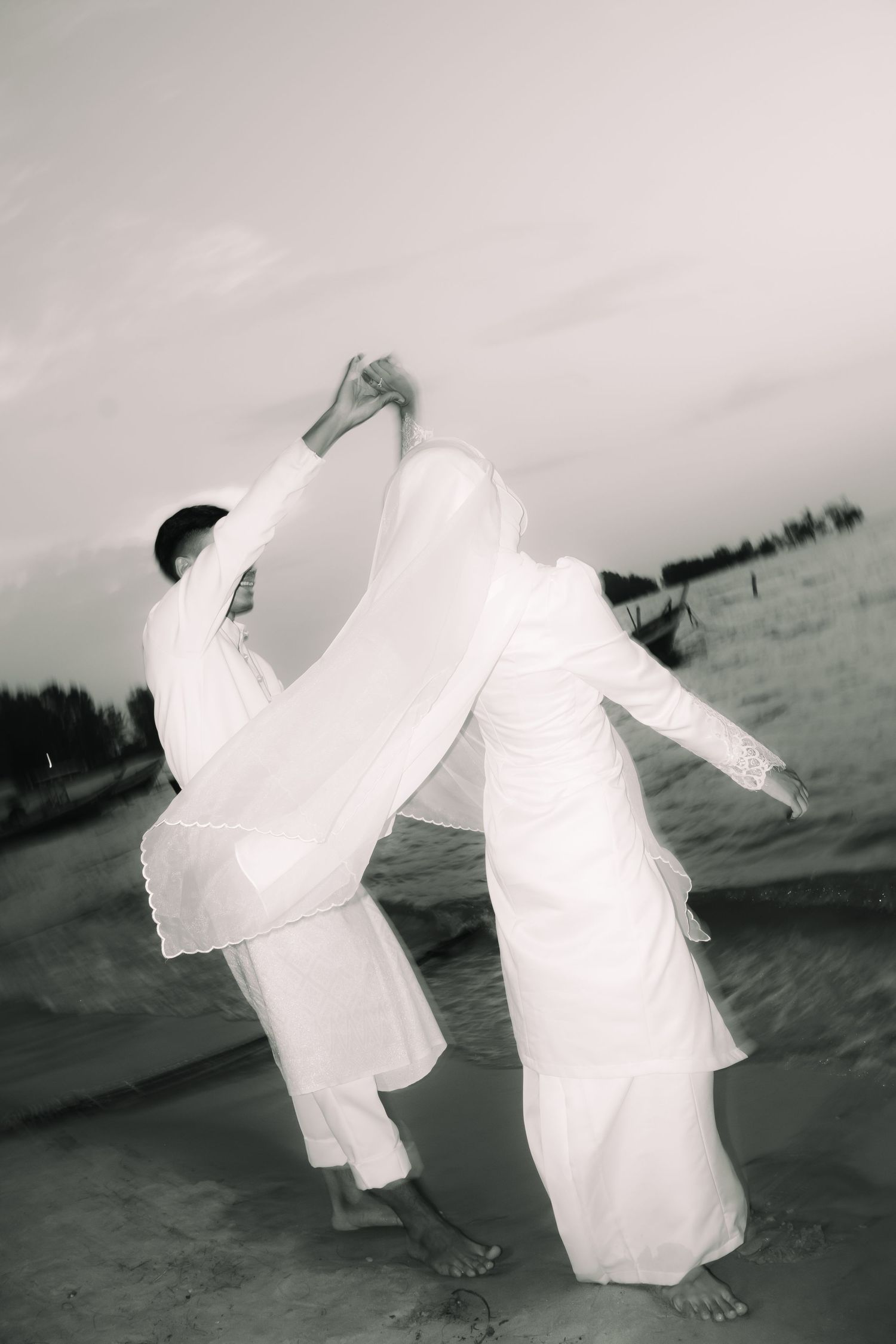 Two figures in flowing white attire perform an artistic dance movement on a beachfront in black and white photography.