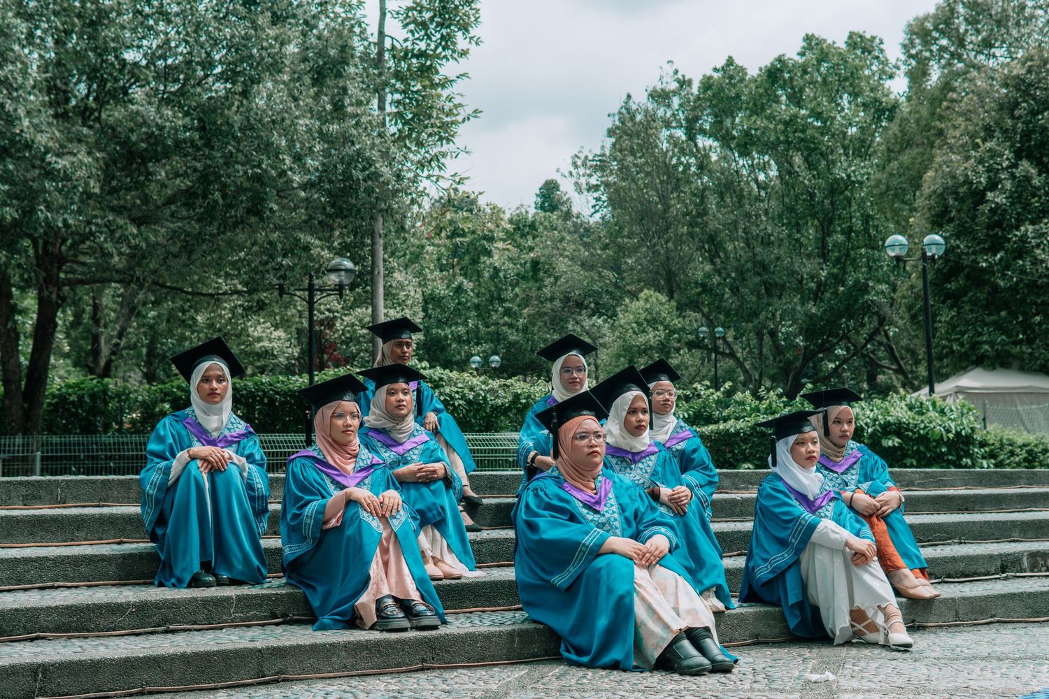 Graduates in blue academic regalia sitting on outdoor amphitheater steps.