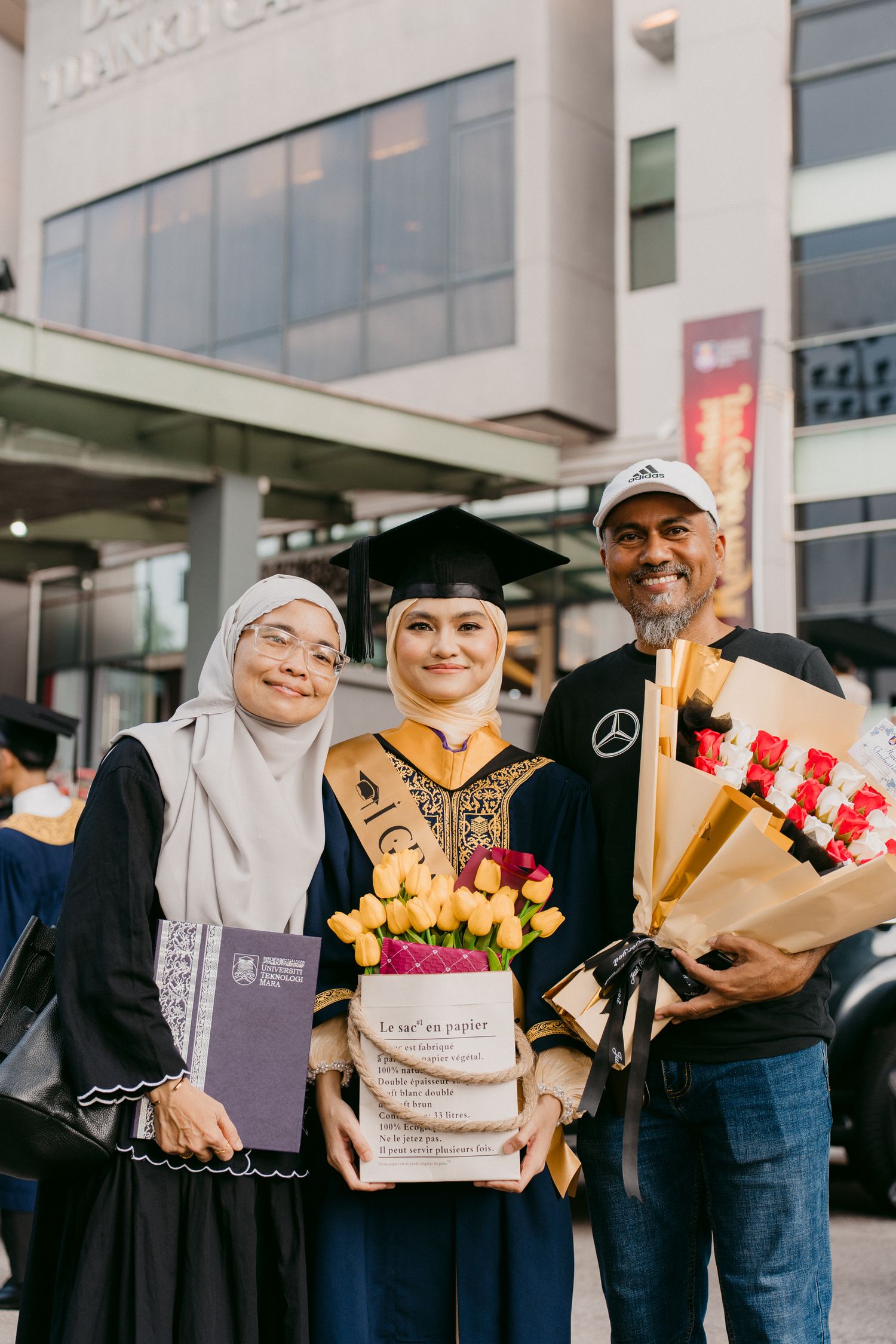 A graduate celebrates with two family members holding flowers and diploma outside a university building.