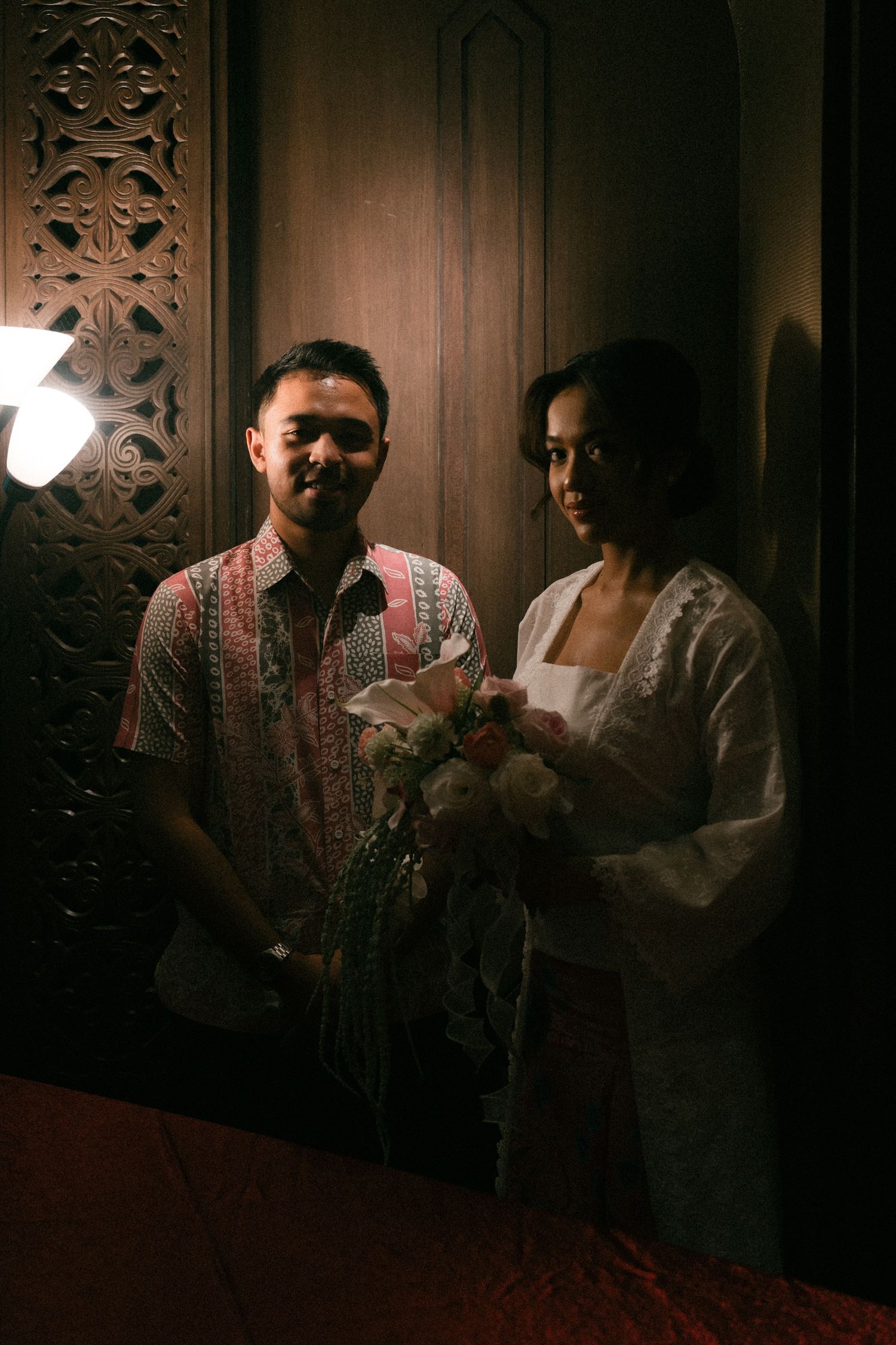 A couple stands in a dimly lit room with ornate wooden wall carvings, captured in moody atmospheric lighting.