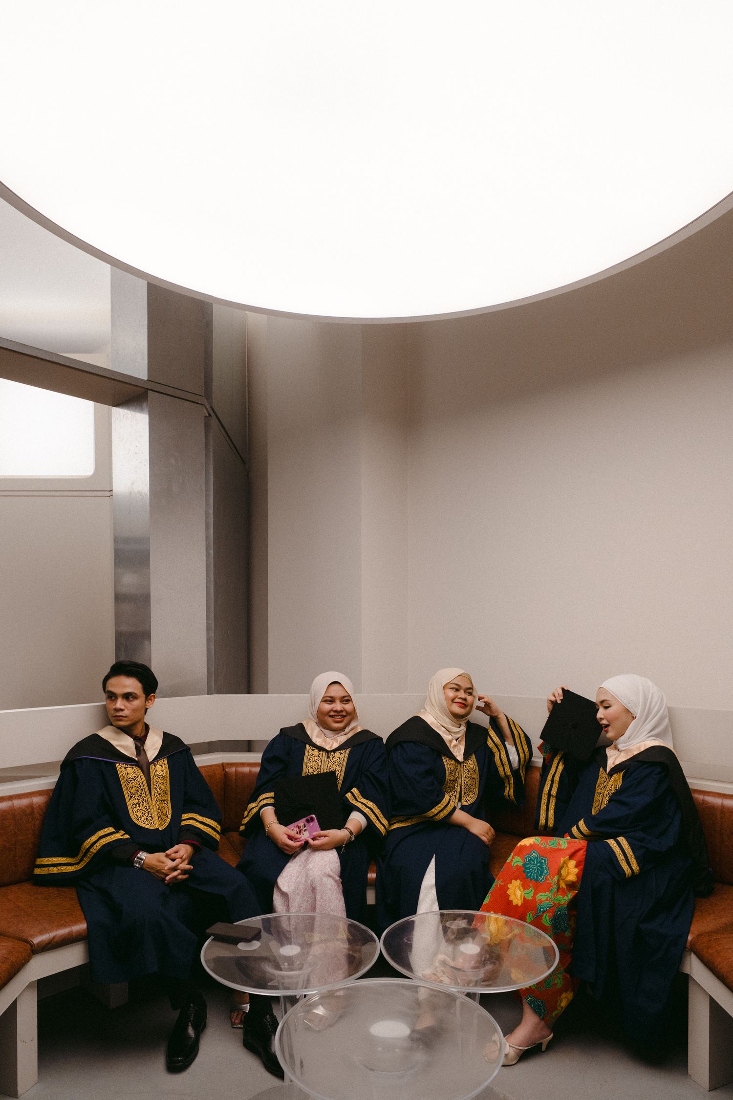Group wearing matching black and gold traditional graduation robes sitting together in a modern room.