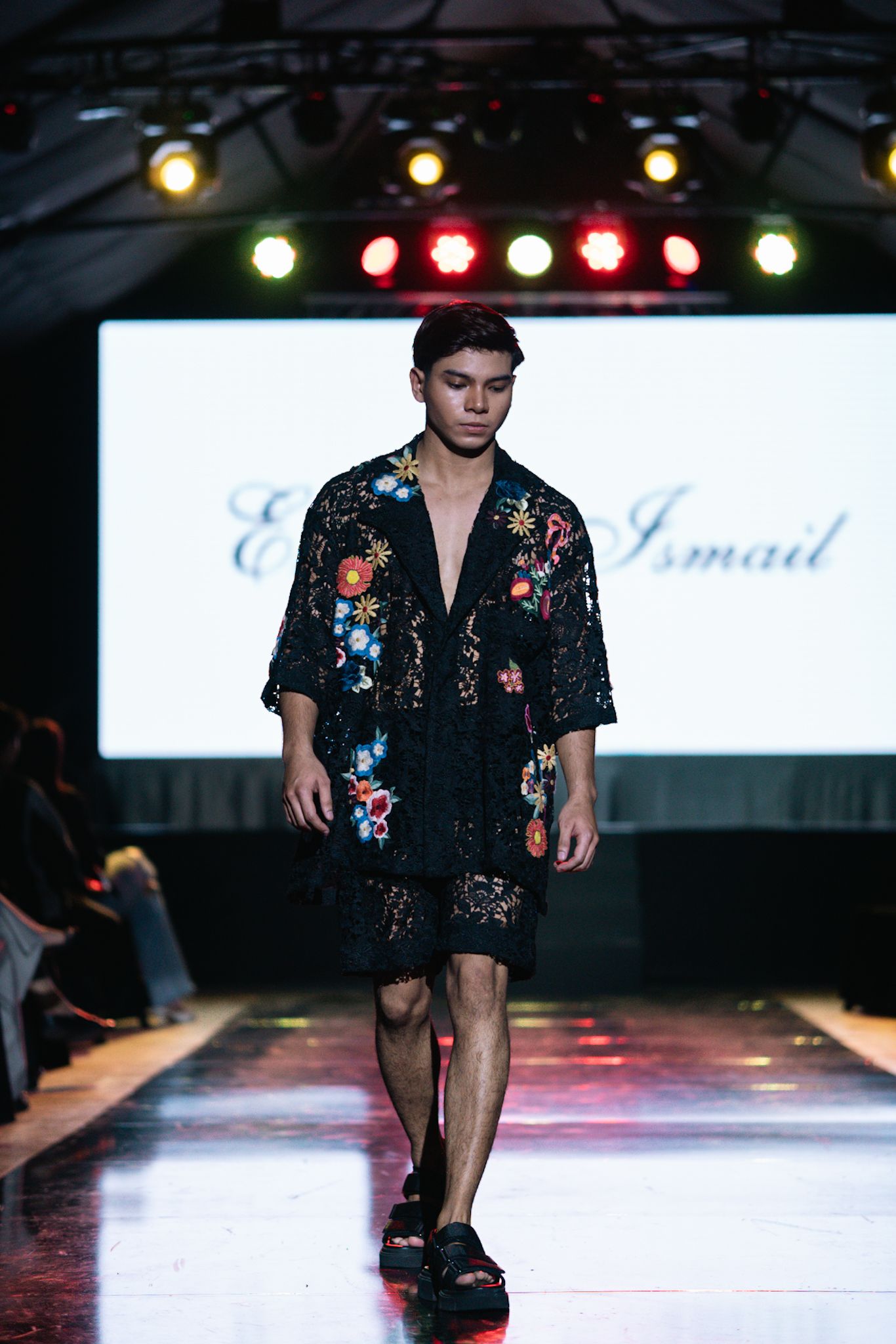 Model in black floral embroidered kimono-style outfit walks fashion runway.