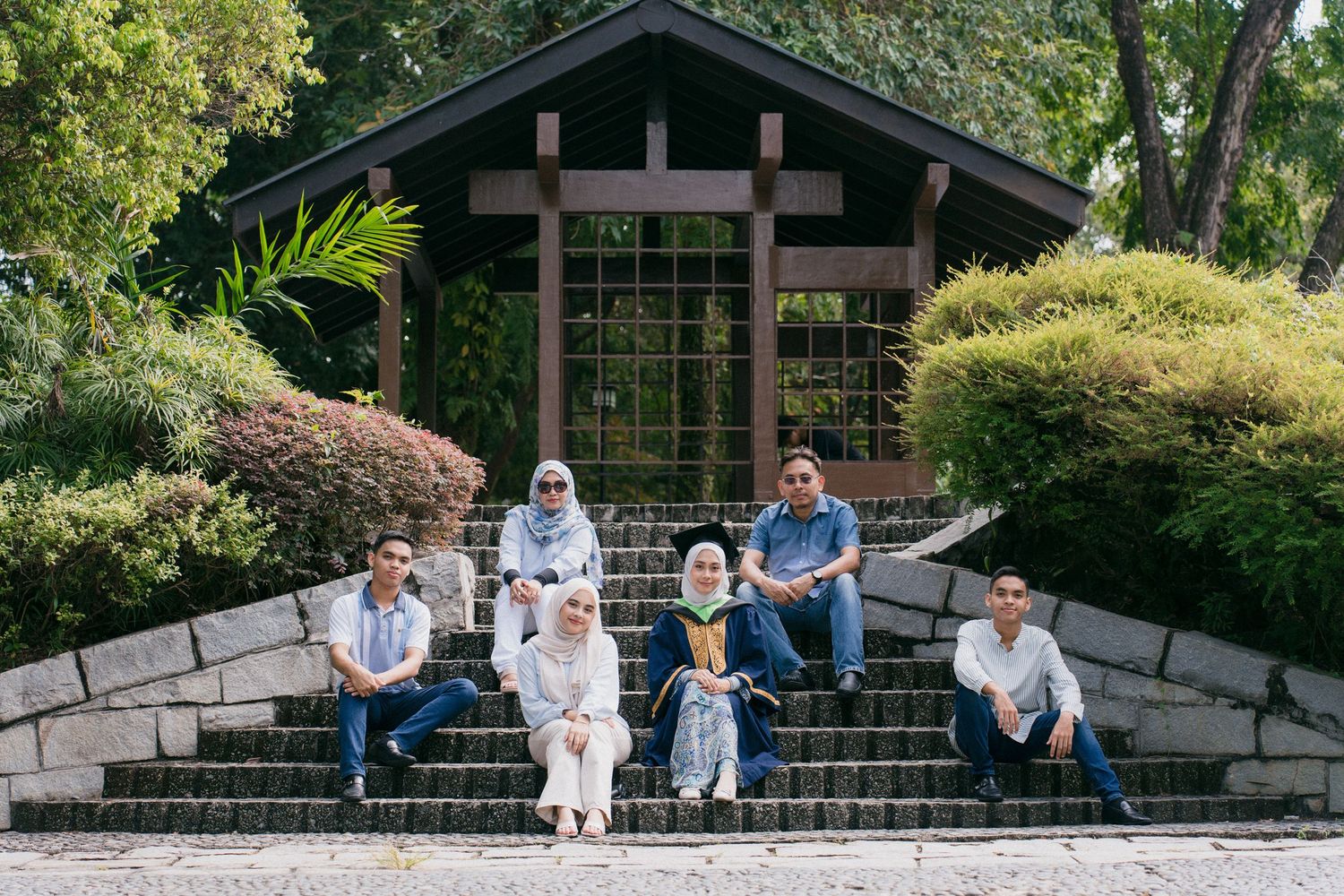 A group sits together on stone steps in a garden setting with greenery and architectural elements in the background.