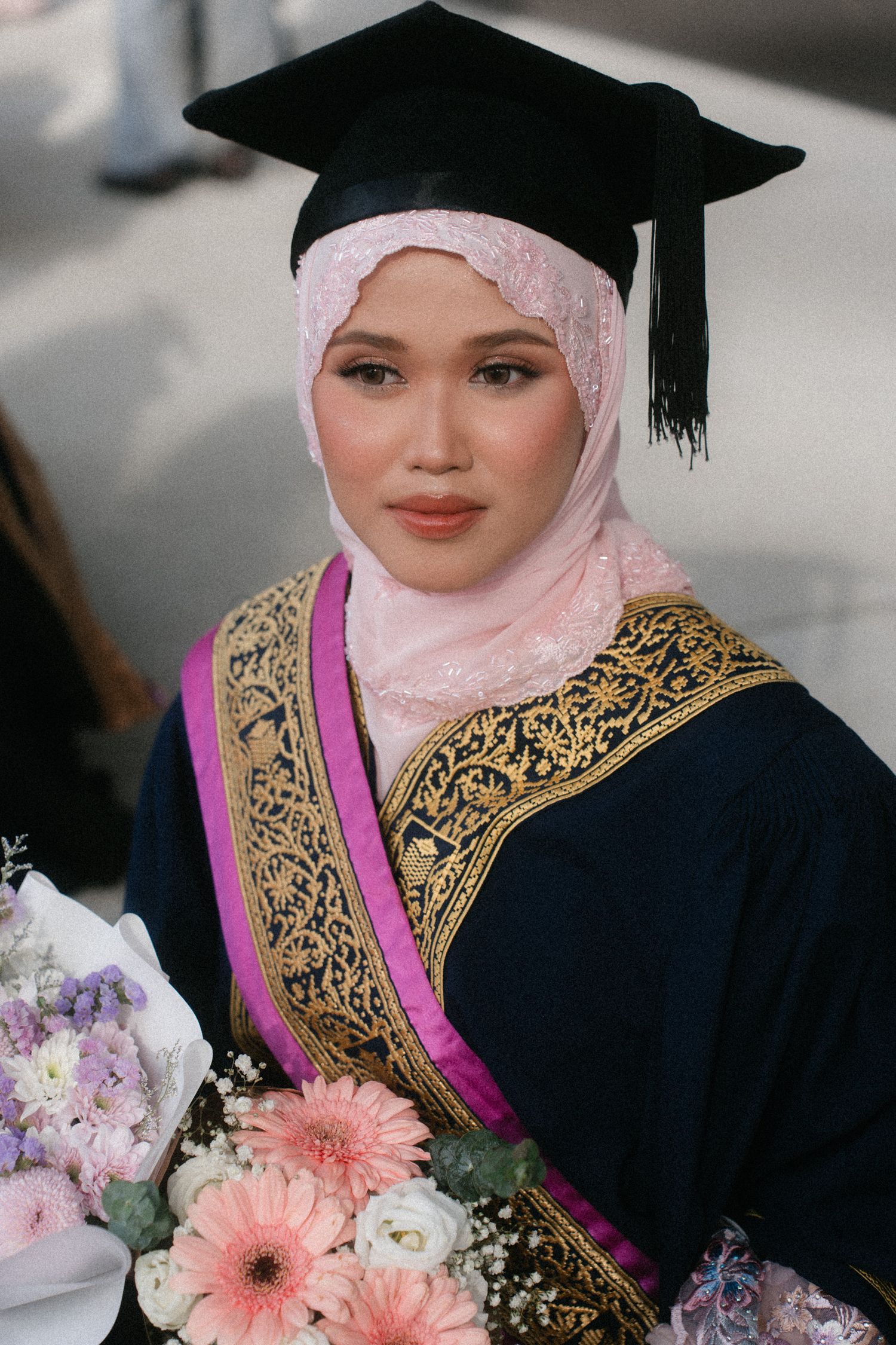 Graduate in black academic regalia with pink hijab and gold embroidered sash holding flowers at commencement ceremony.