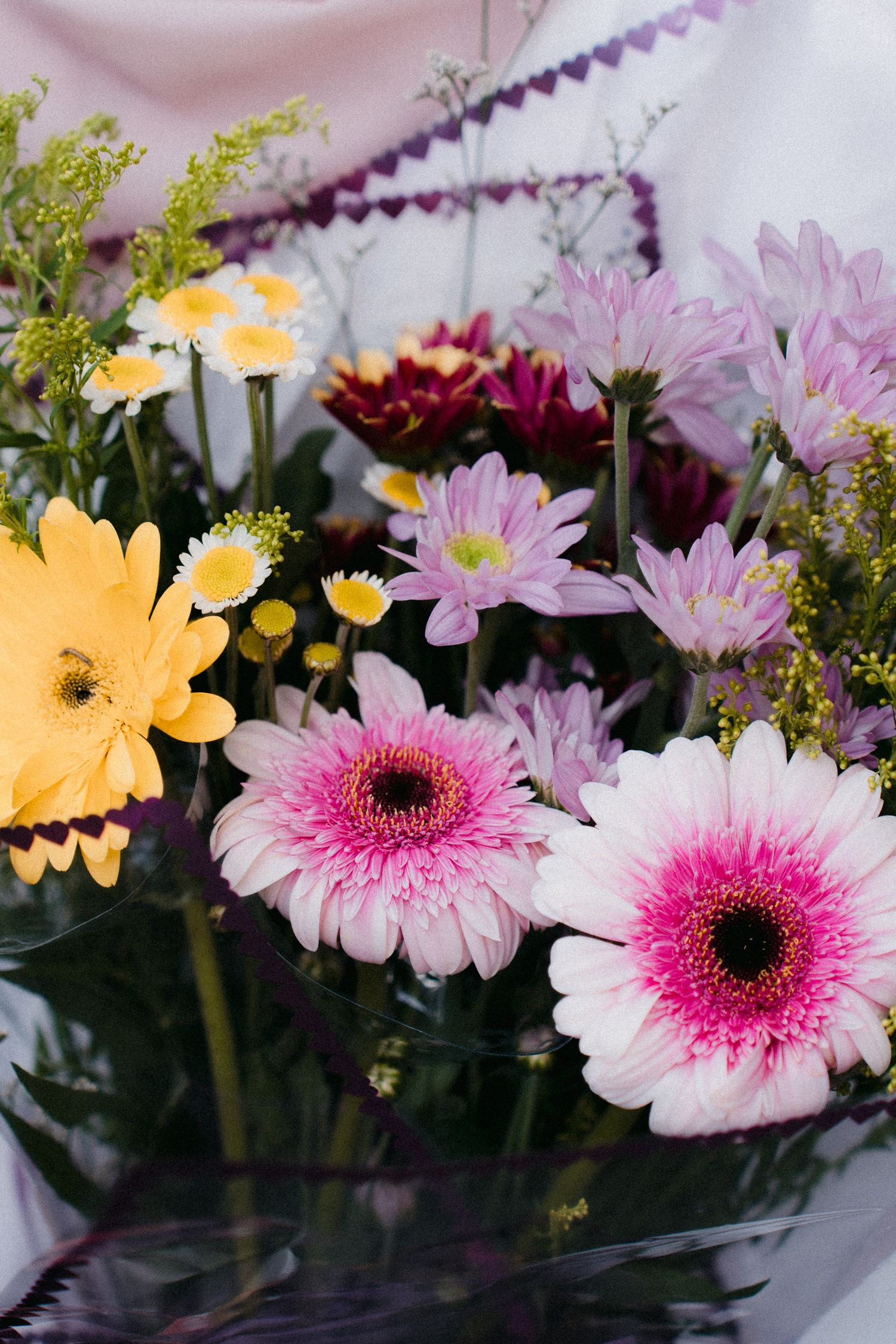 A colorful flower arrangement featuring pink gerbera daisies, yellow daisies, and delicate purple flowers in a vase.