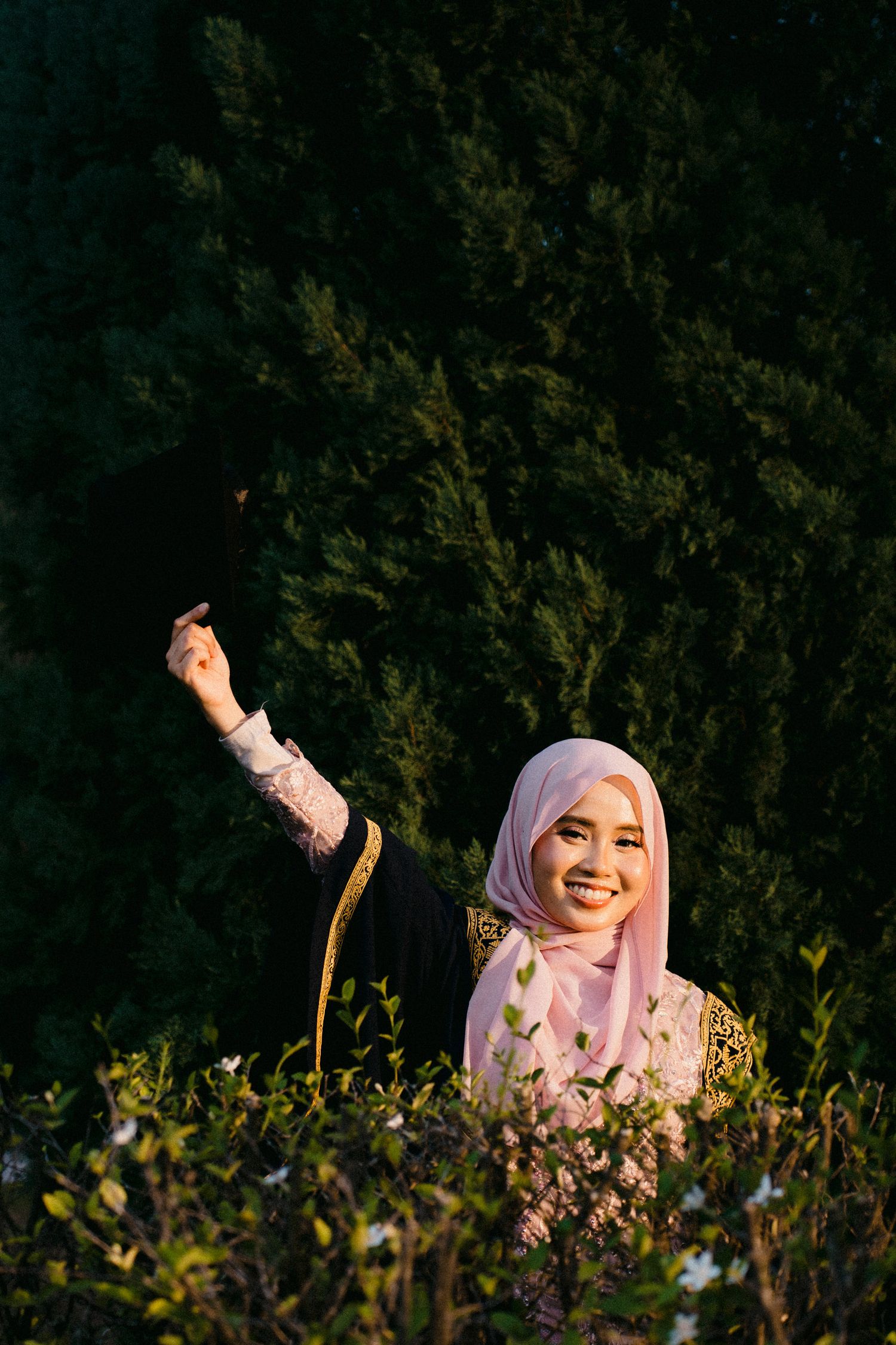 A person in a pink hijab smiles and poses among green foliage in a series of outdoor portrait shots.