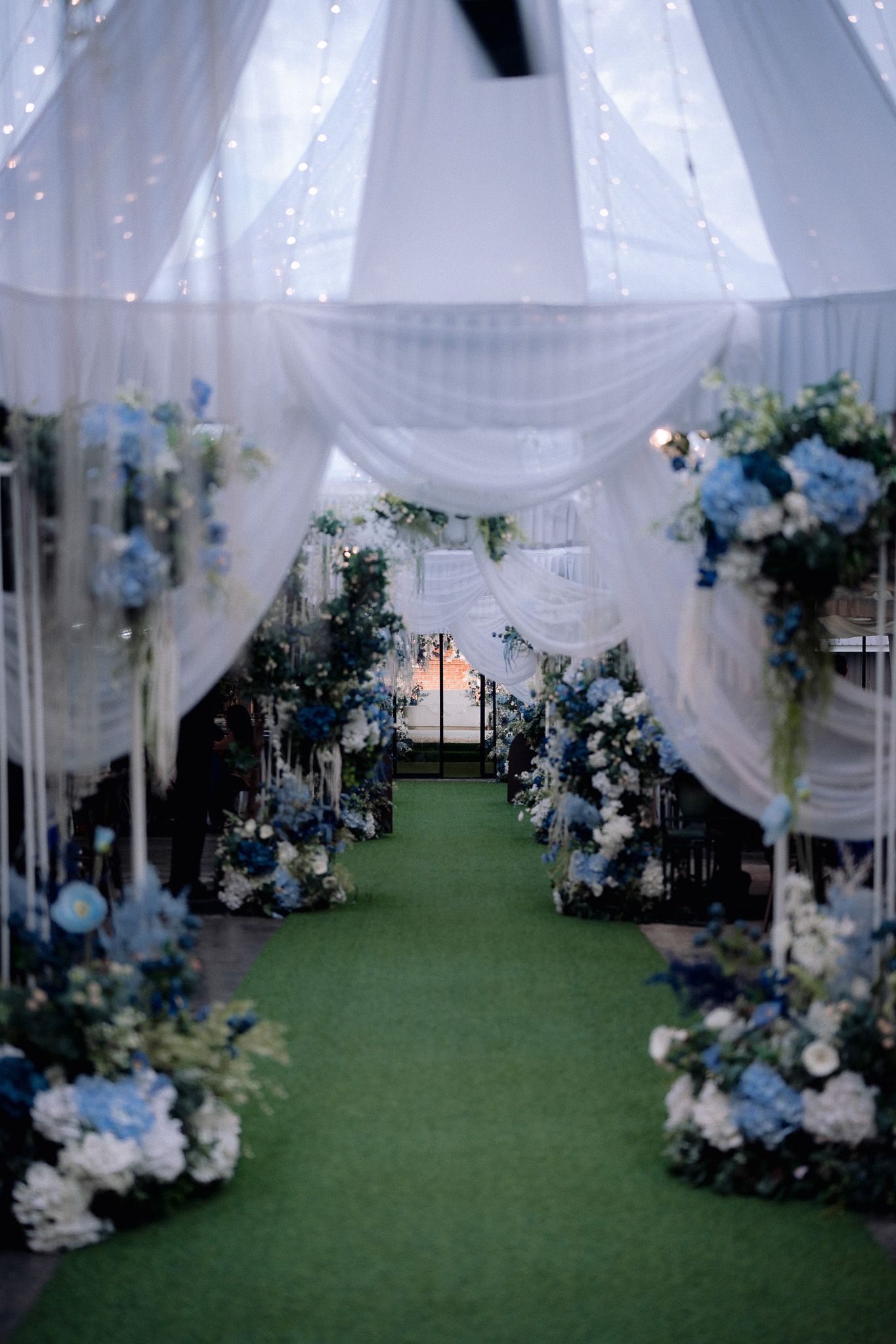 Elegant wedding aisle with blue and white floral arrangements, green carpet, and white draped fabric canopy with fairy lights.