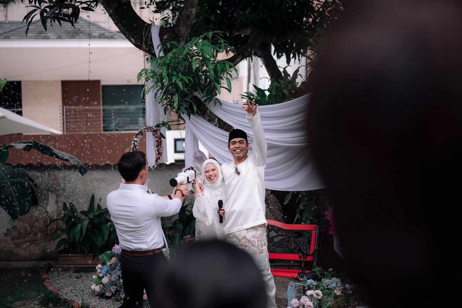 A couple in white wedding attire celebrates outdoors as a photographer captures their joyful moment during an Islamic wedding ceremony.