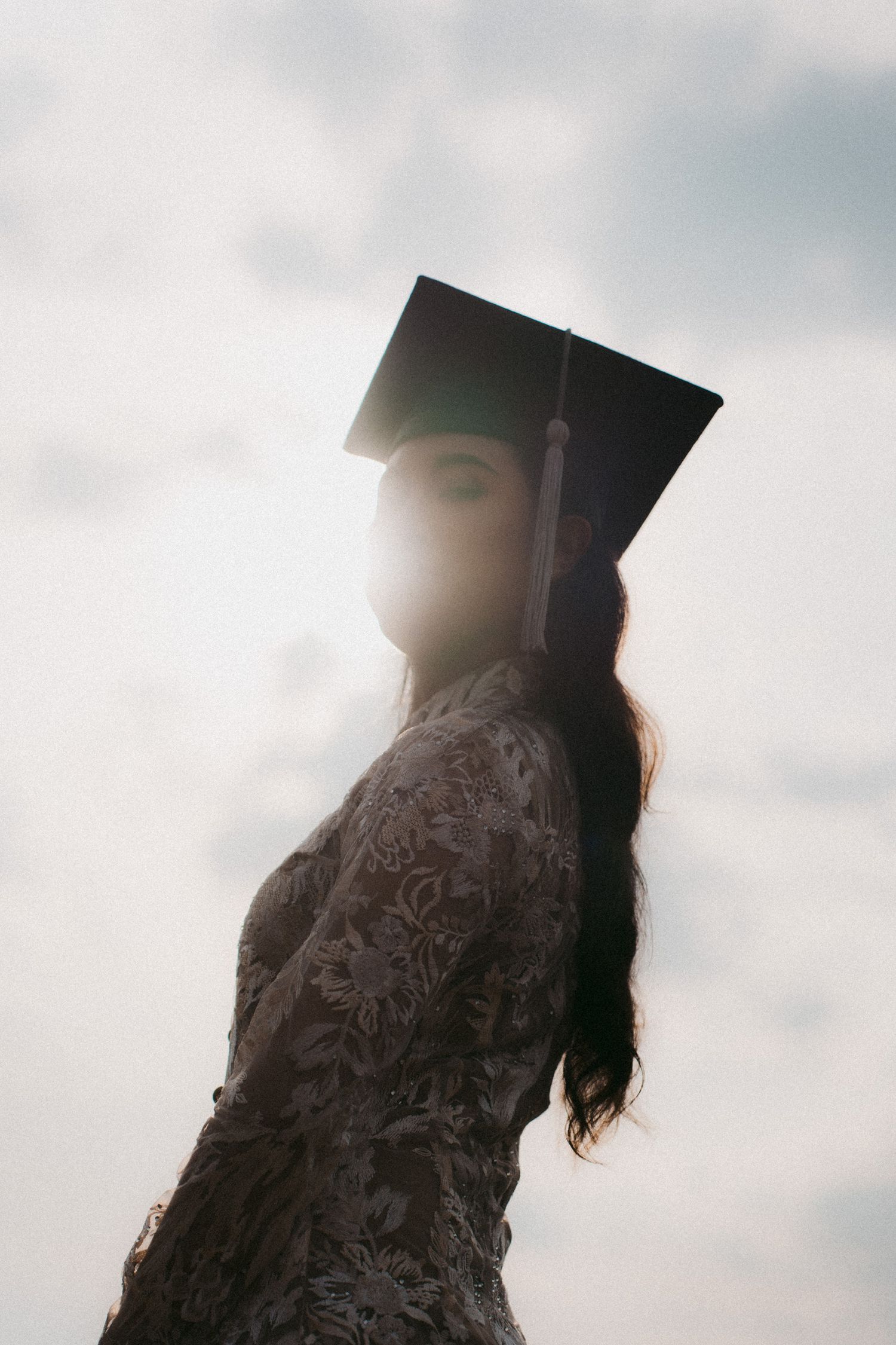 A graduate in cap and gown stands in silhouette against a cloudy sky during a graduation ceremony.