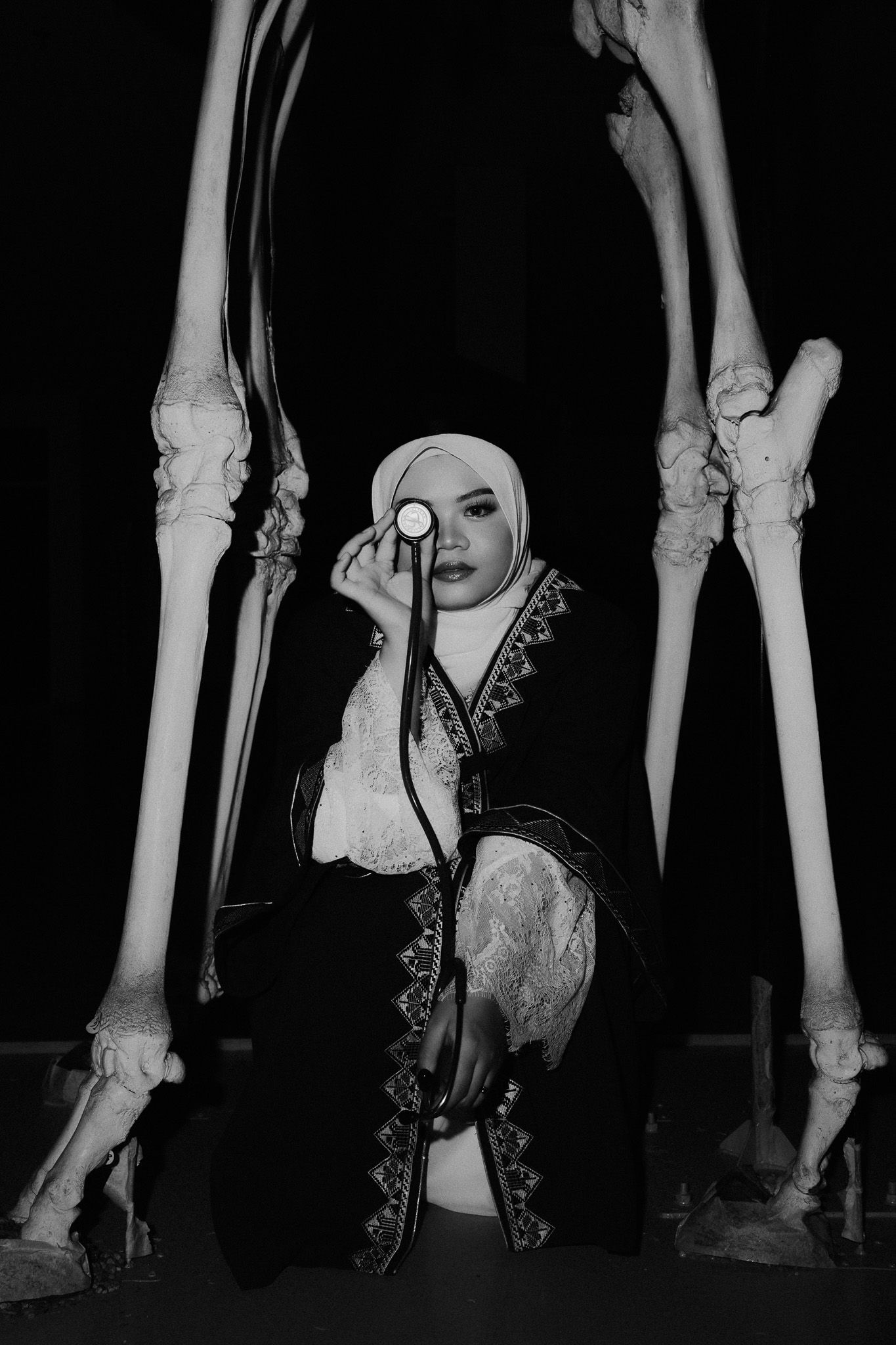 Black and white photo of a woman in hijab and embroidered robe holding a monocle, framed by large animal bones.