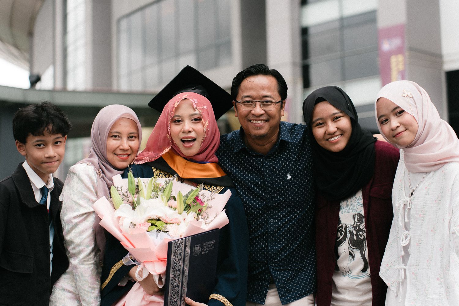 A graduate in cap and gown holds flowers surrounded by smiling family members at a graduation ceremony.