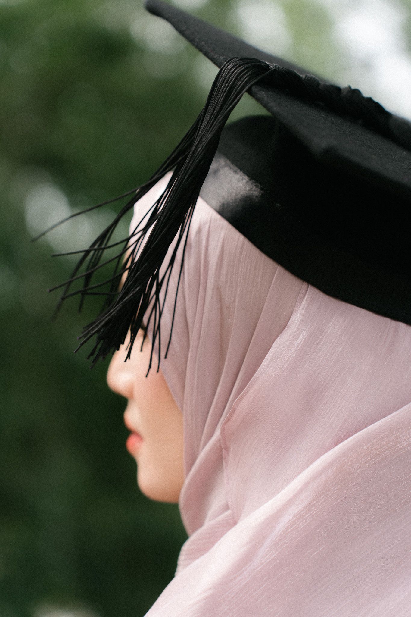 A Muslim woman in a pink hijab wearing a black graduation mortarboard cap, photographed in profile outdoors.