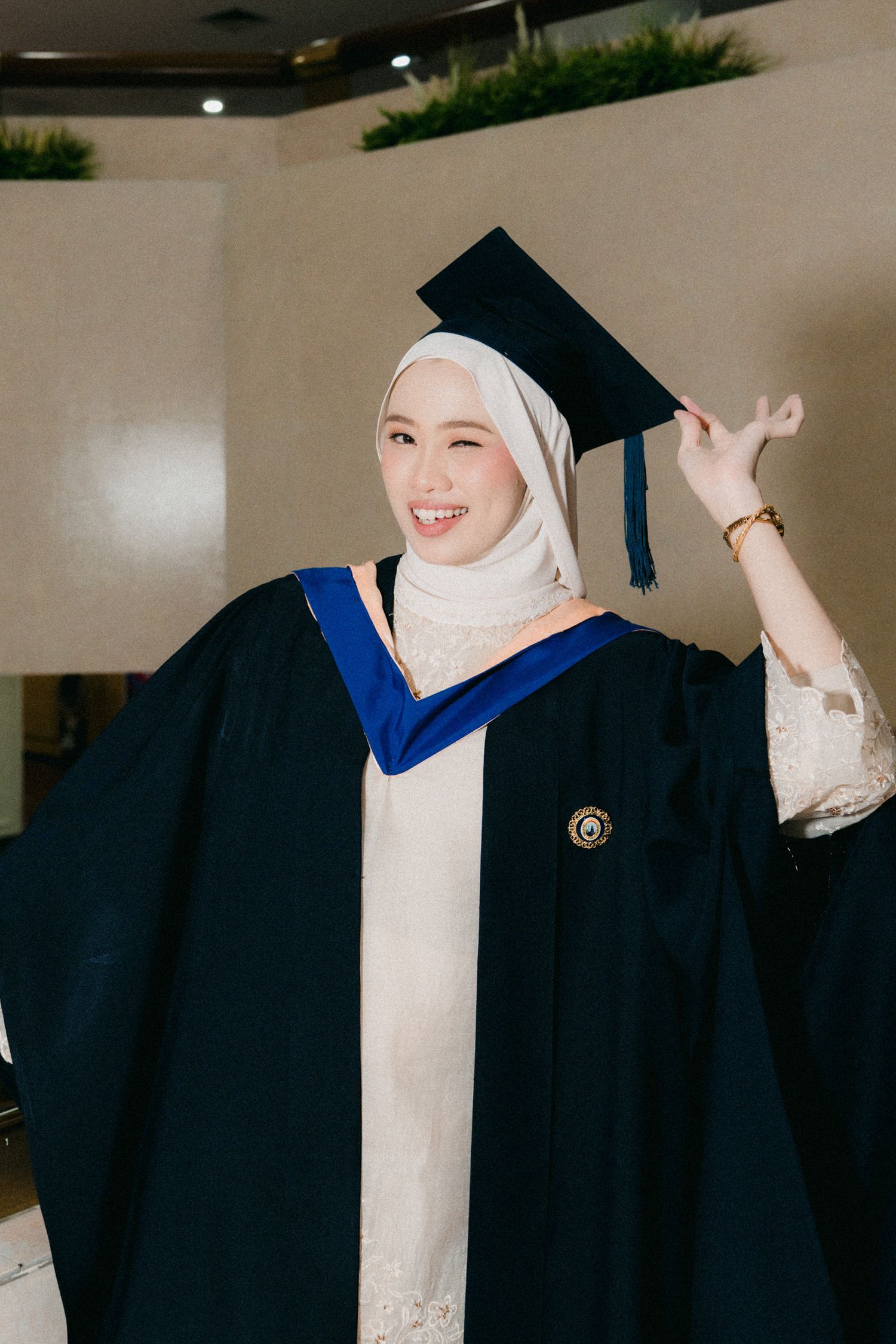 A graduate in traditional academic regalia and white hijab poses joyfully during commencement ceremony.