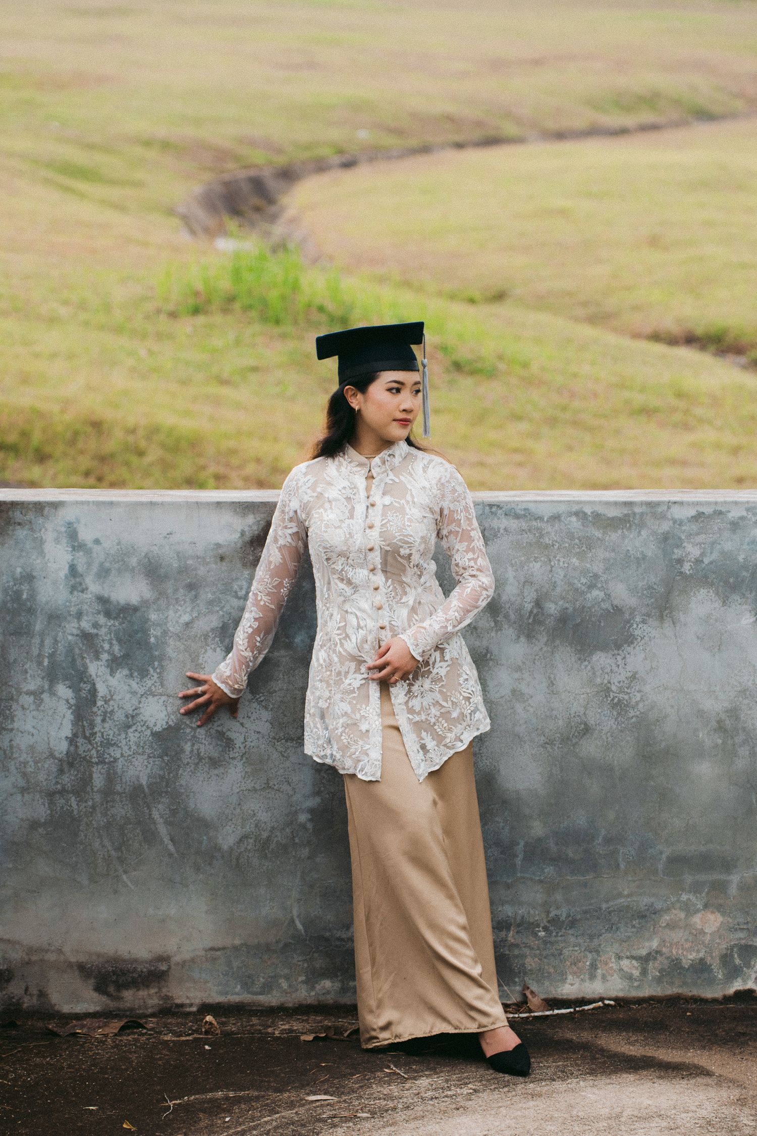 A person in a white lace top and beige skirt poses against a concrete wall in a grassy outdoor setting.