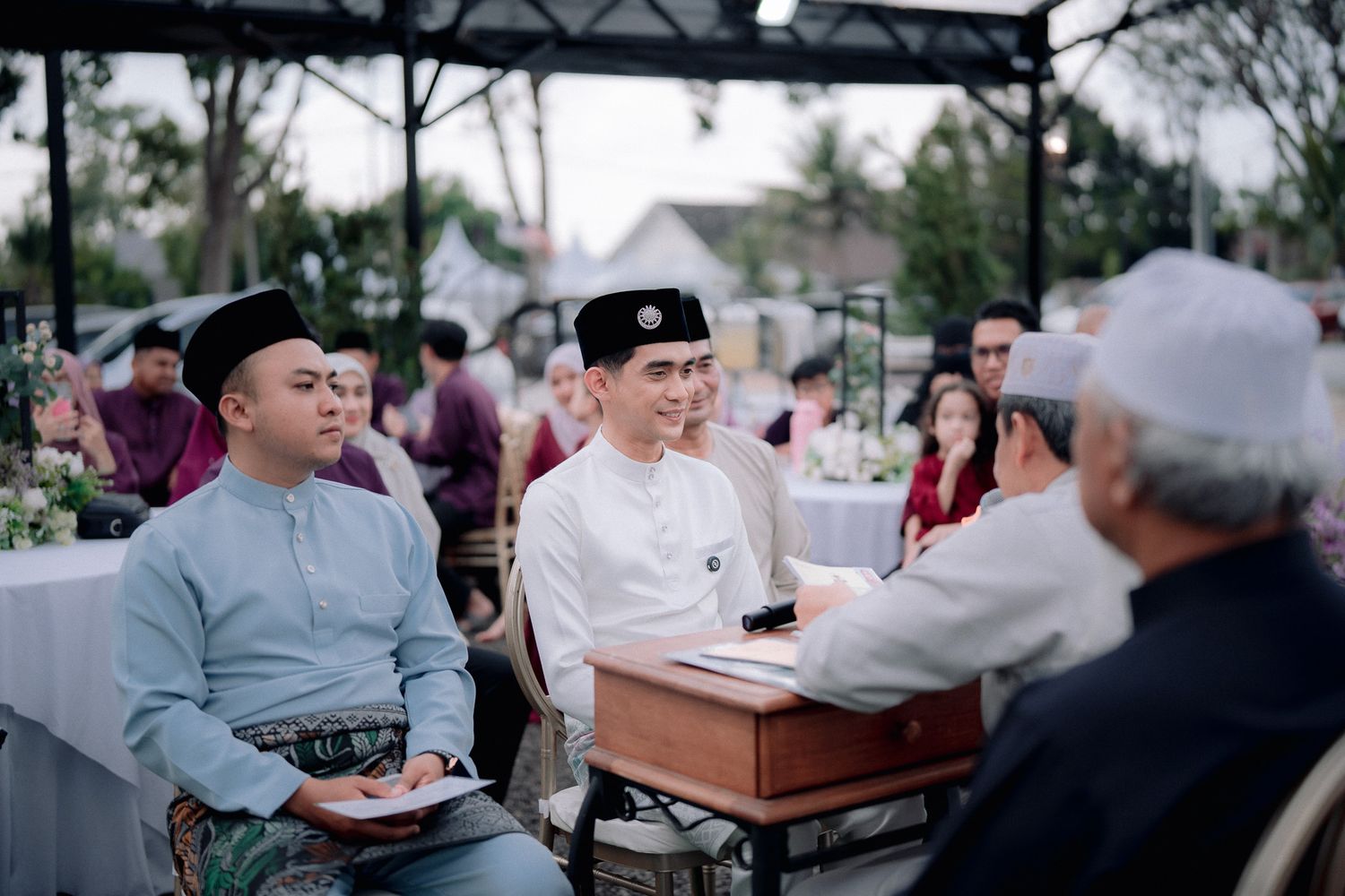 Men in traditional Malay attire socializing at an outdoor wedding reception event.