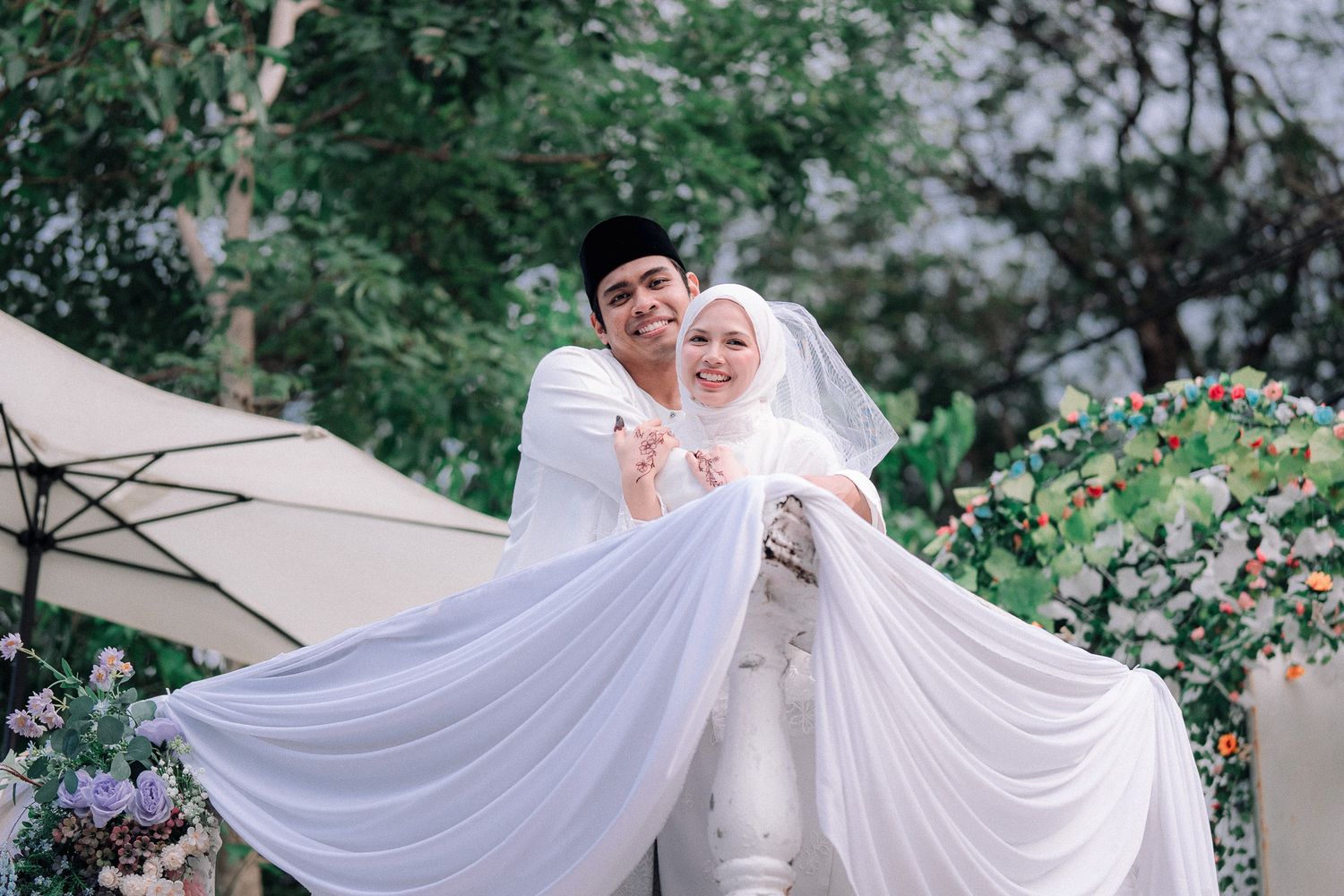 Muslim couple in white wedding attire posing joyfully outdoors, bride in hijab and groom lifting her veil, surrounded by flowers.