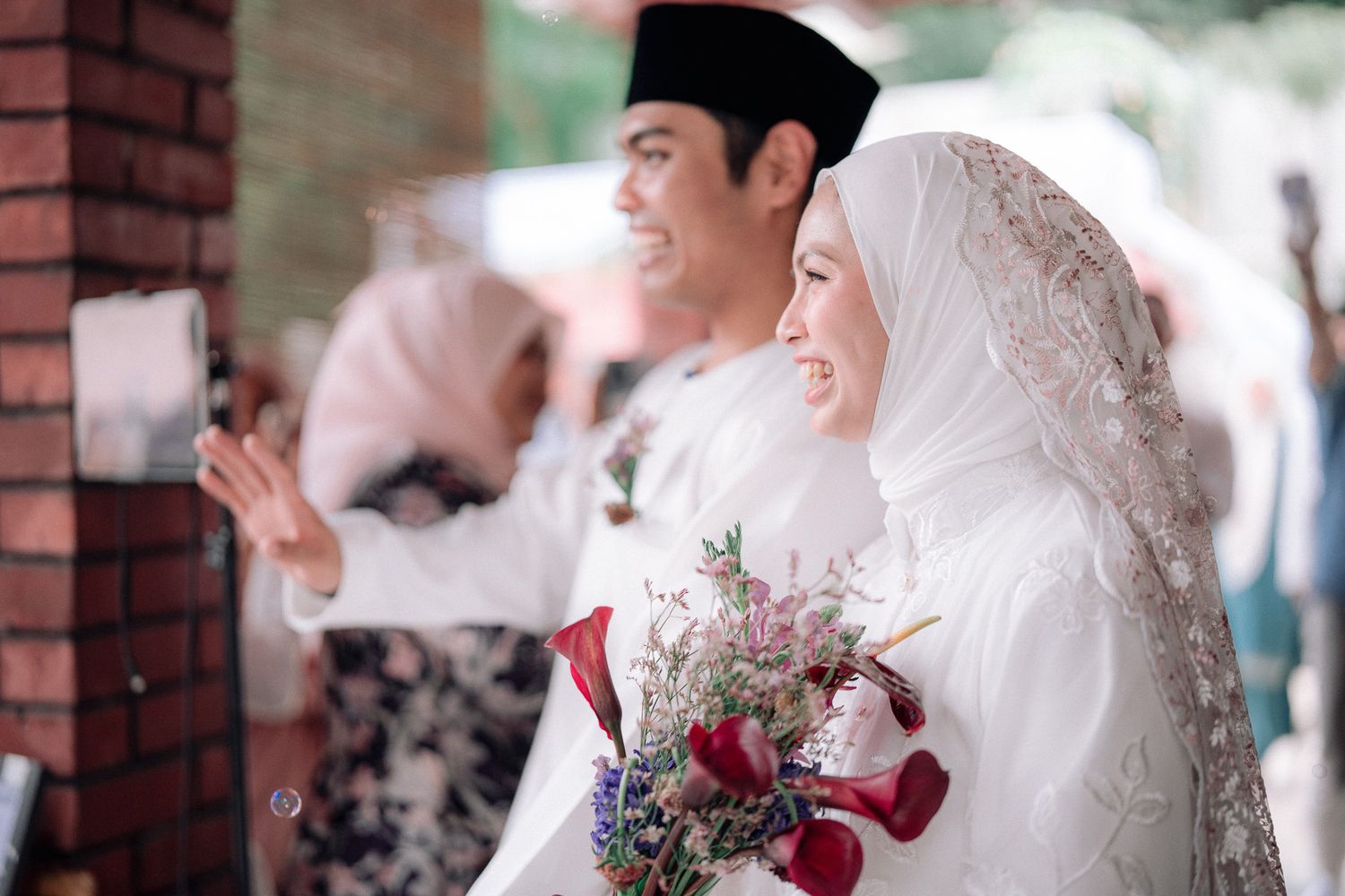 Muslim couple in traditional wedding attire, bride holding floral bouquet in white hijab and groom in white baju melayu with songkok.