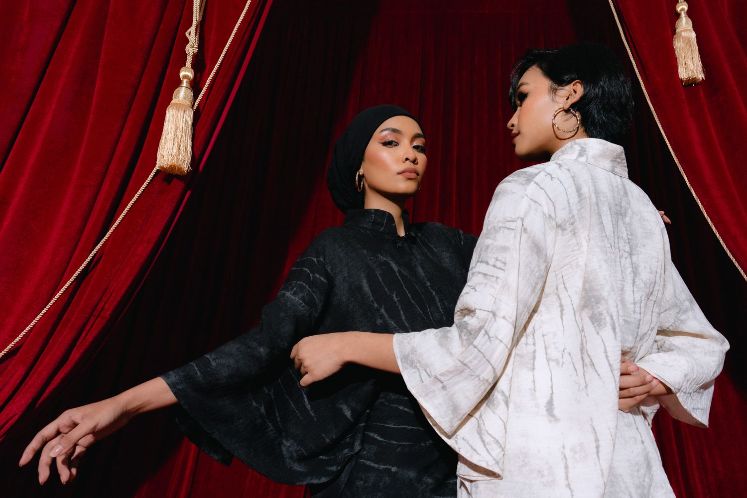 Two models pose dramatically in black and white garments against rich red velvet curtains in an elegant fashion shoot.