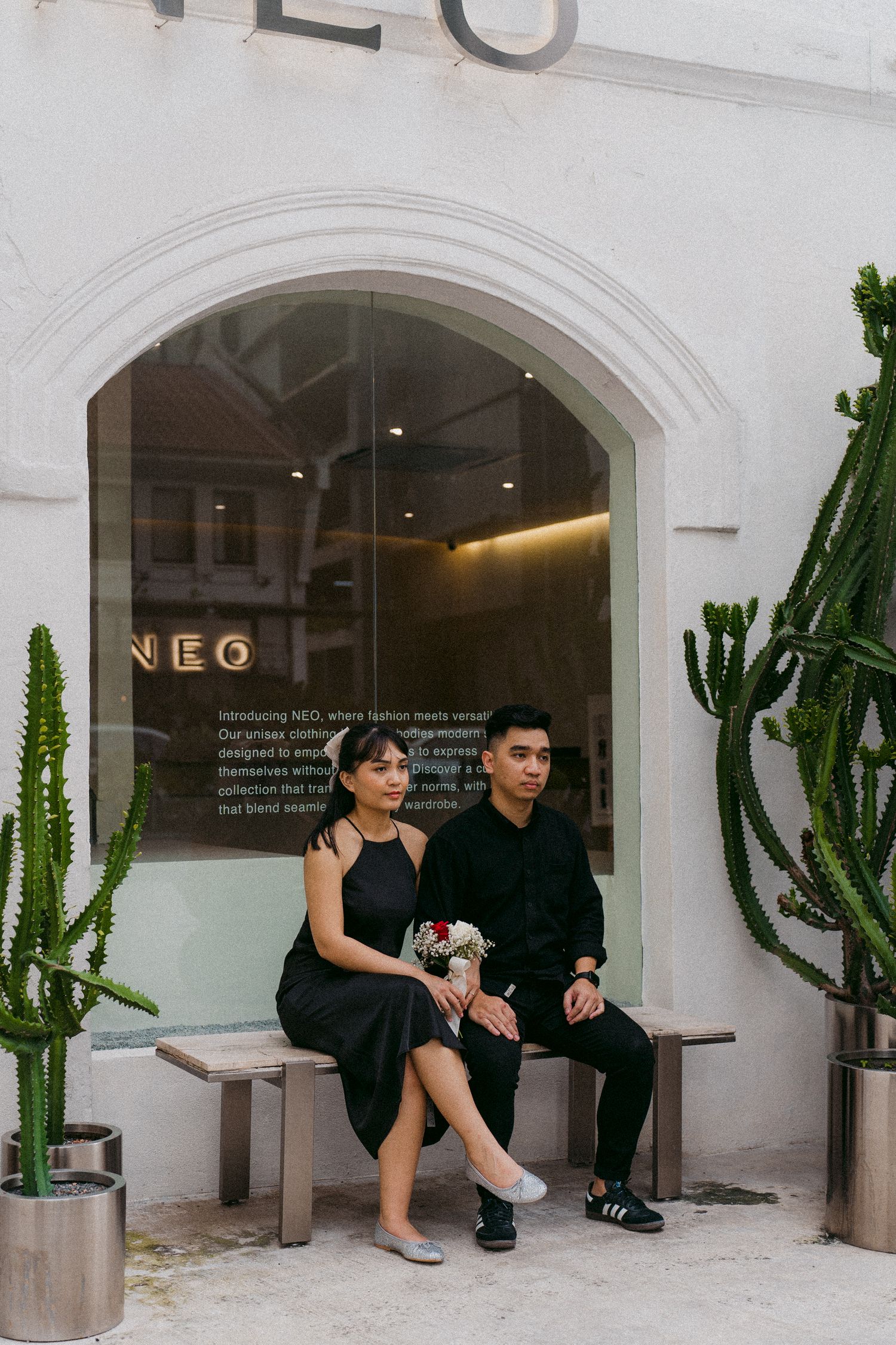 A couple sitting on a bench outside a storefront with NEO signage and decorative cacti.