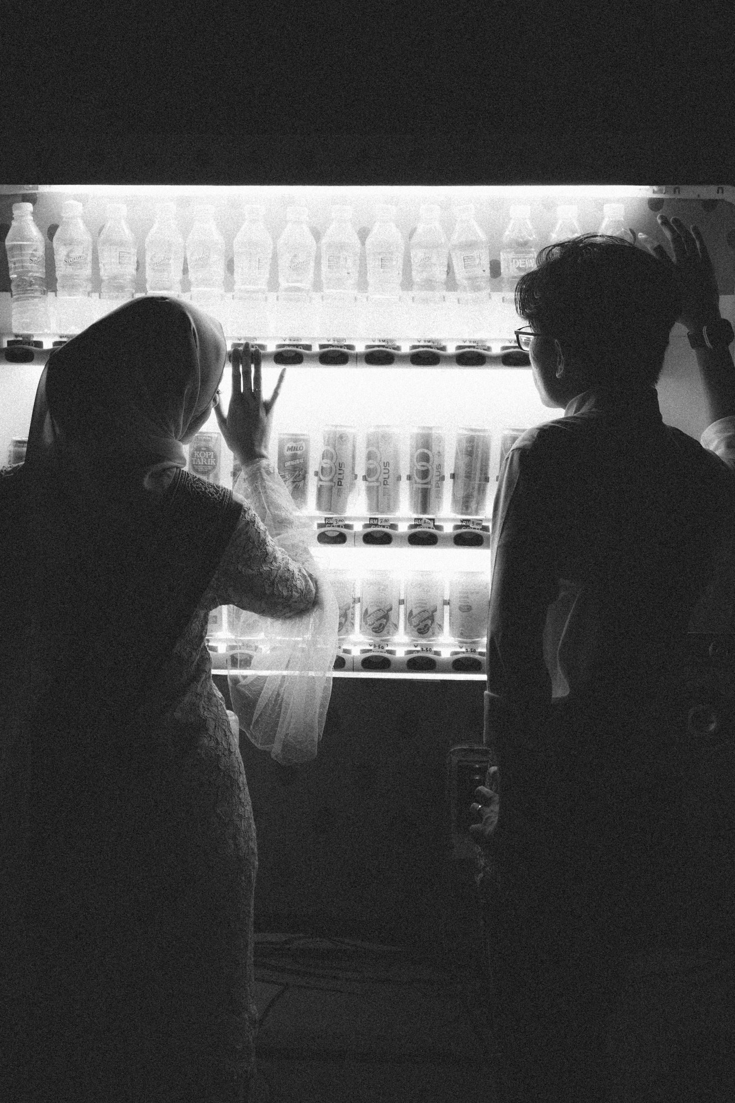 Two silhouettes examine illuminated bottles on display shelves.