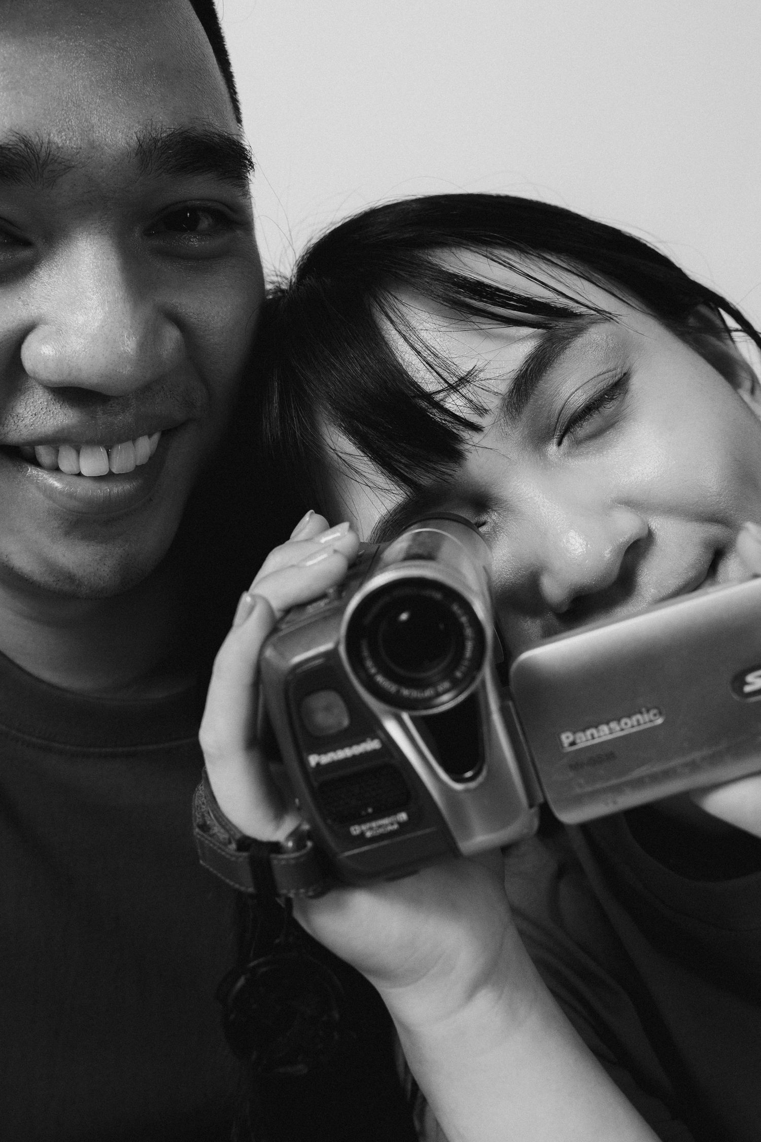 Couple smiling while holding vintage camcorder in black and white photography.