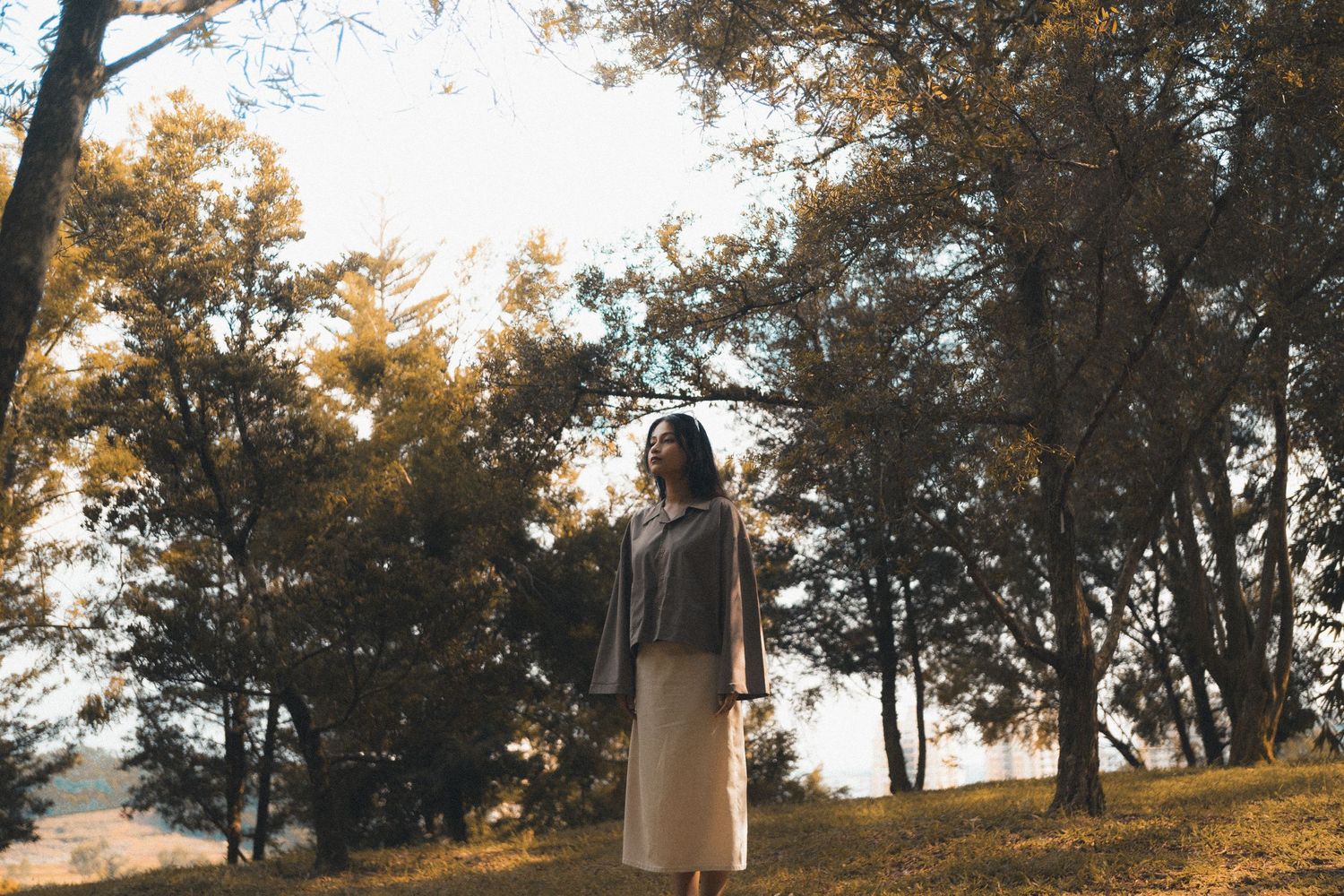 Figure in light clothing stands on hillside surrounded by trees in warm sunlight.