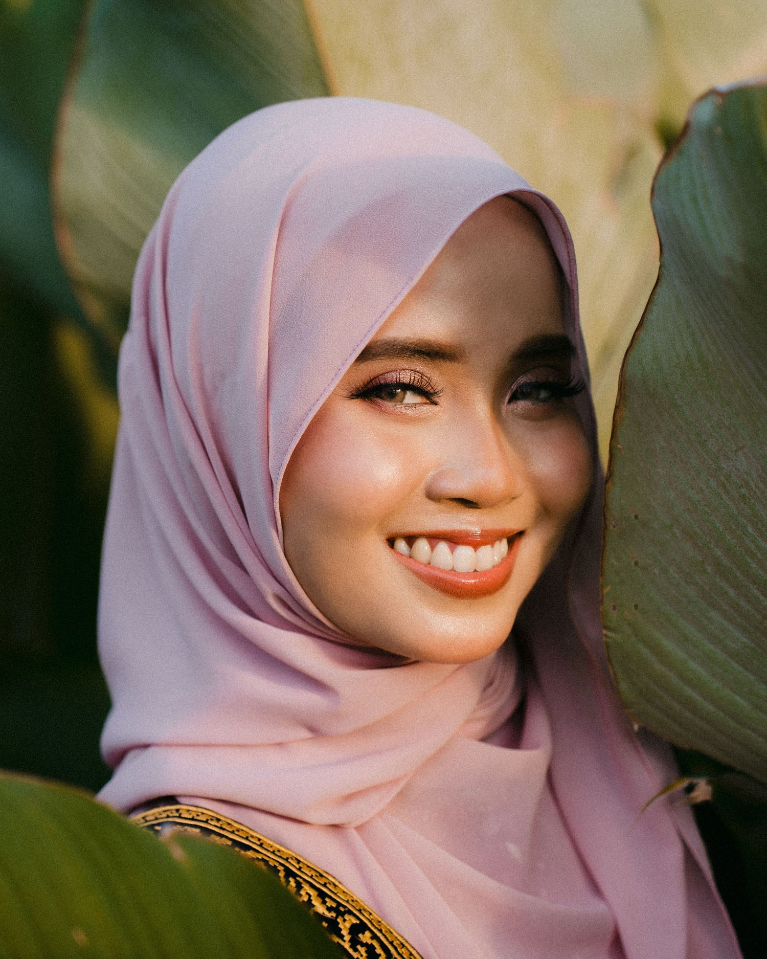 A woman in a light pink hijab smiles warmly while posing against a blurred natural background.