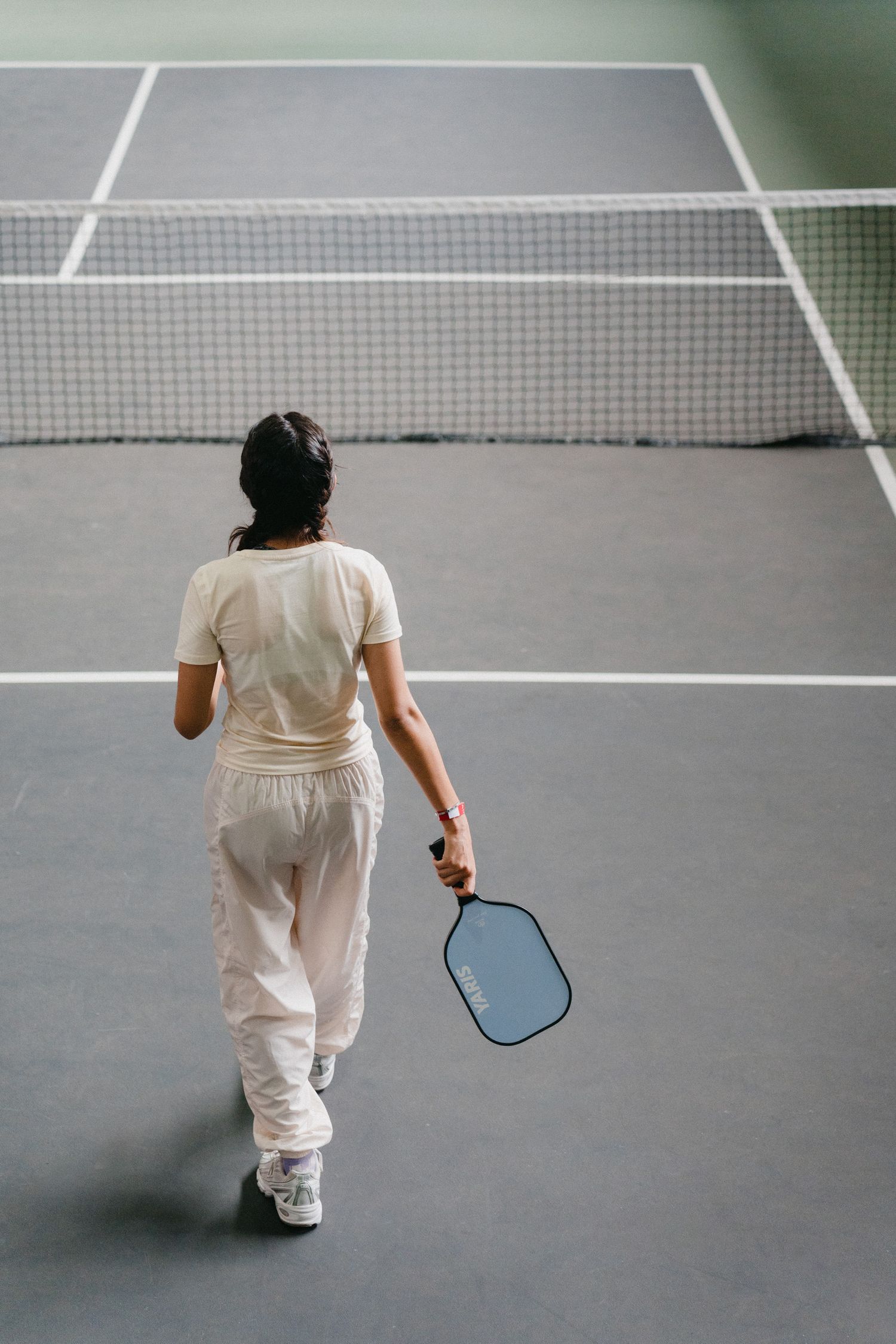 Tennis player in white attire walking across an indoor court with a racquet, viewed from behind.