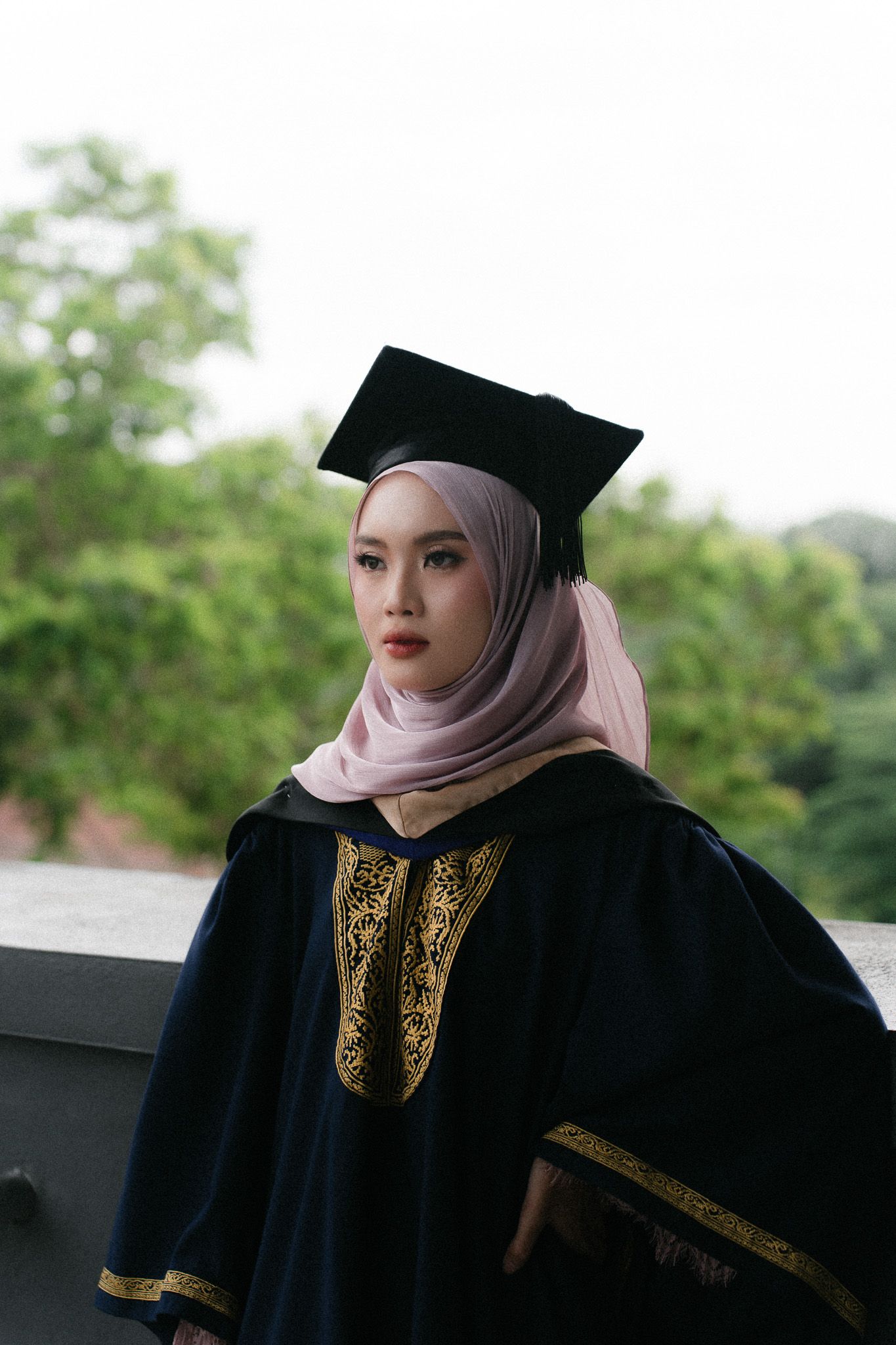 Young Muslim woman in graduation gown and mortarboard cap wearing hijab, posing outdoors with green trees background.