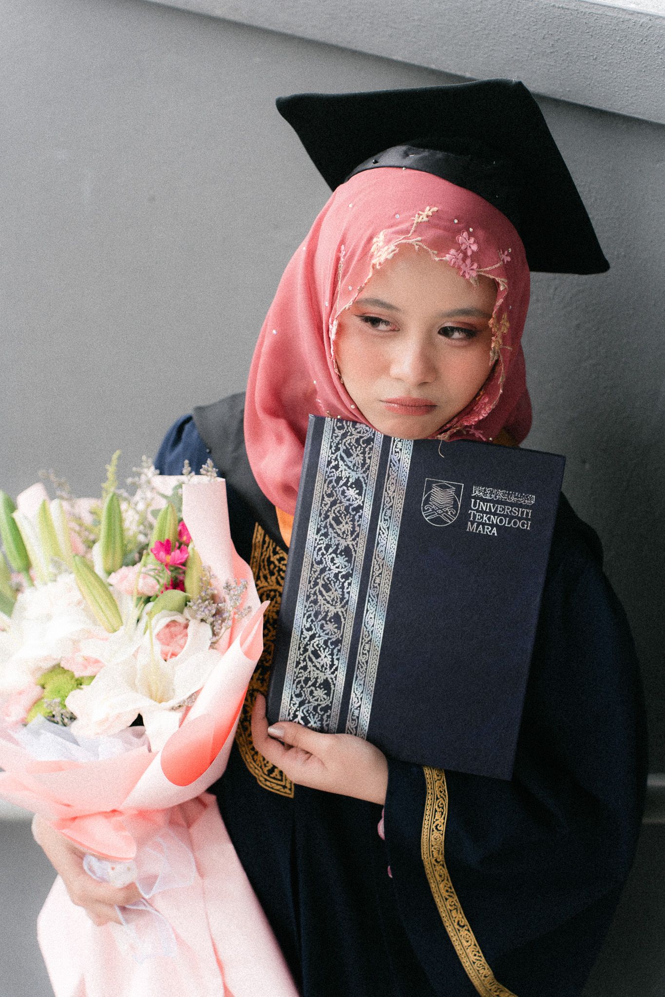 Muslim woman in graduation gown and hijab holding diploma and flowers at university commencement ceremony.