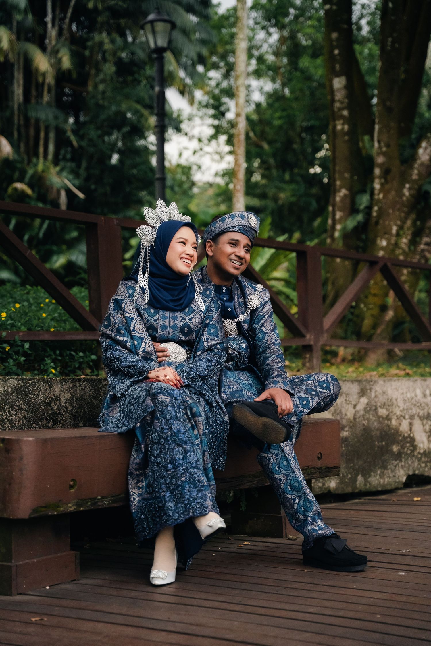 Couple in matching blue floral traditional outfits sitting together on wooden deck surrounded by lush greenery.