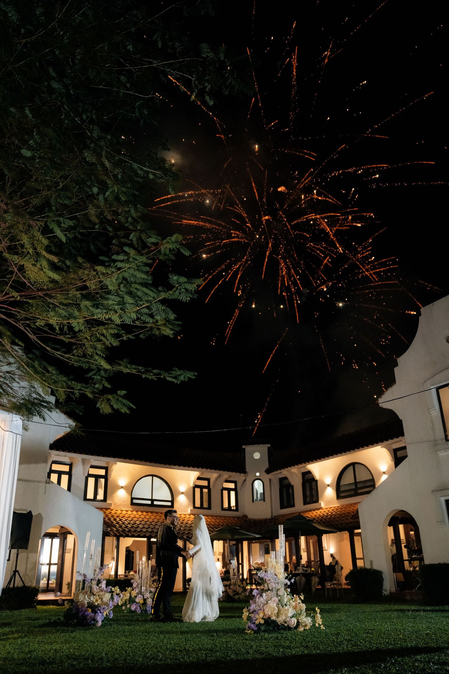 Fireworks light up the night sky over an illuminated white Spanish-style building during an outdoor evening celebration.