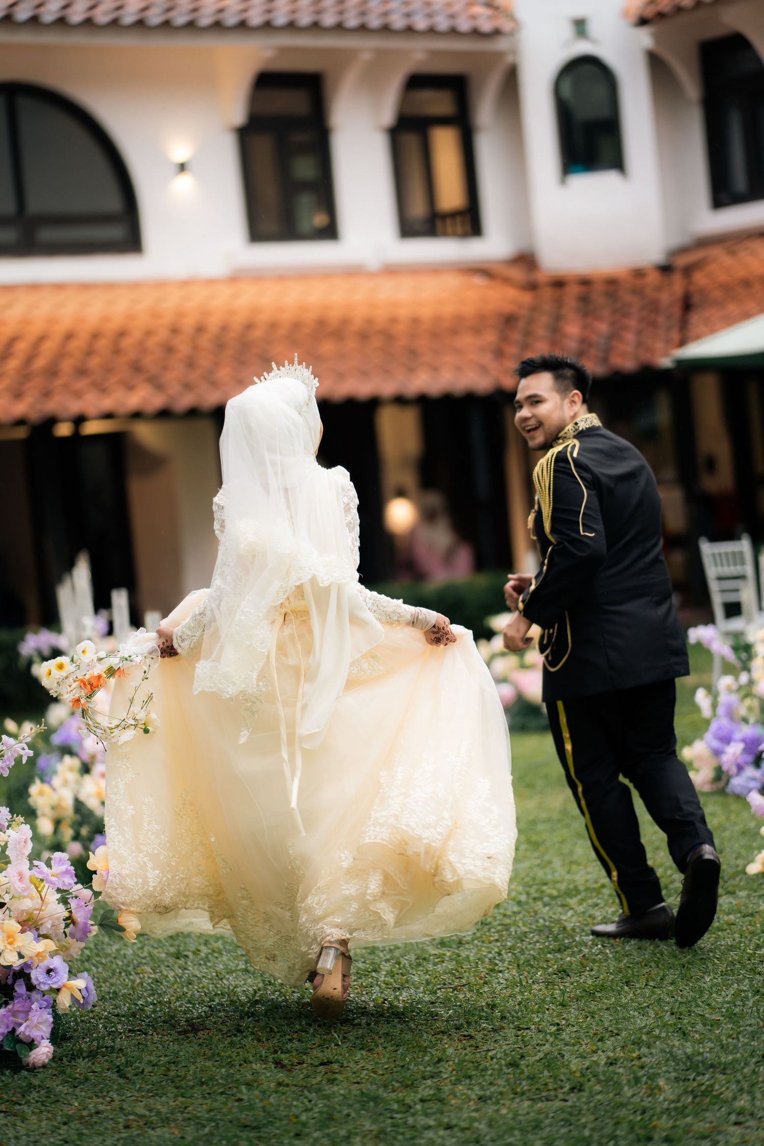 A couple shares a playful moment running on grass with Spanish-style architecture and colorful flowers in the background.