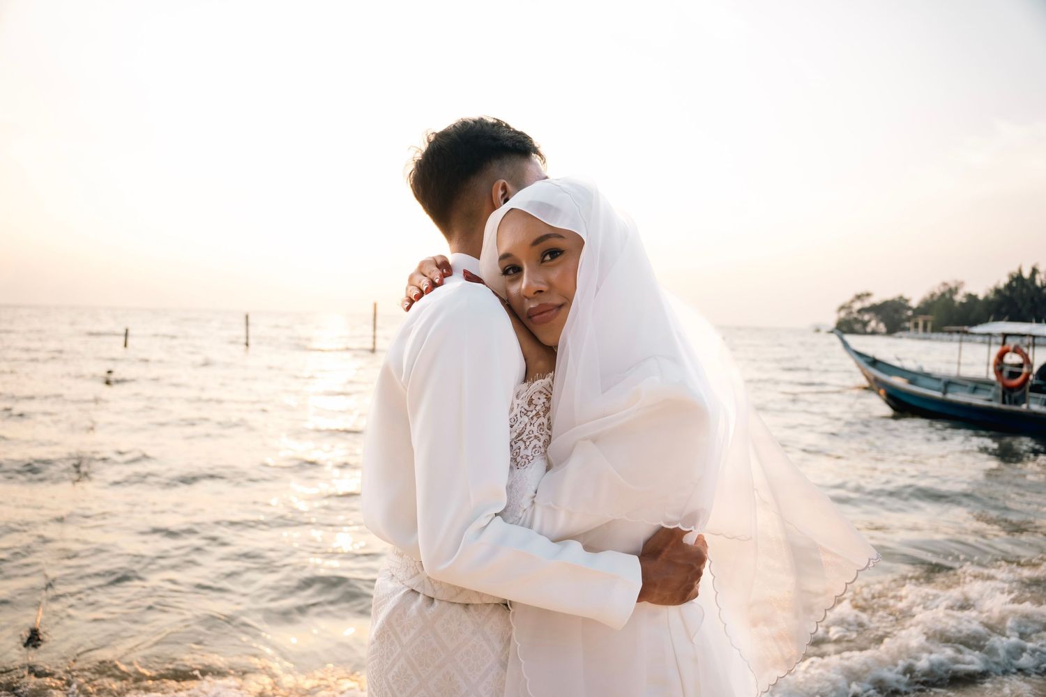 Romantic couple in warm white sweaters embrace by the ocean during golden hour as waves crash behind them.