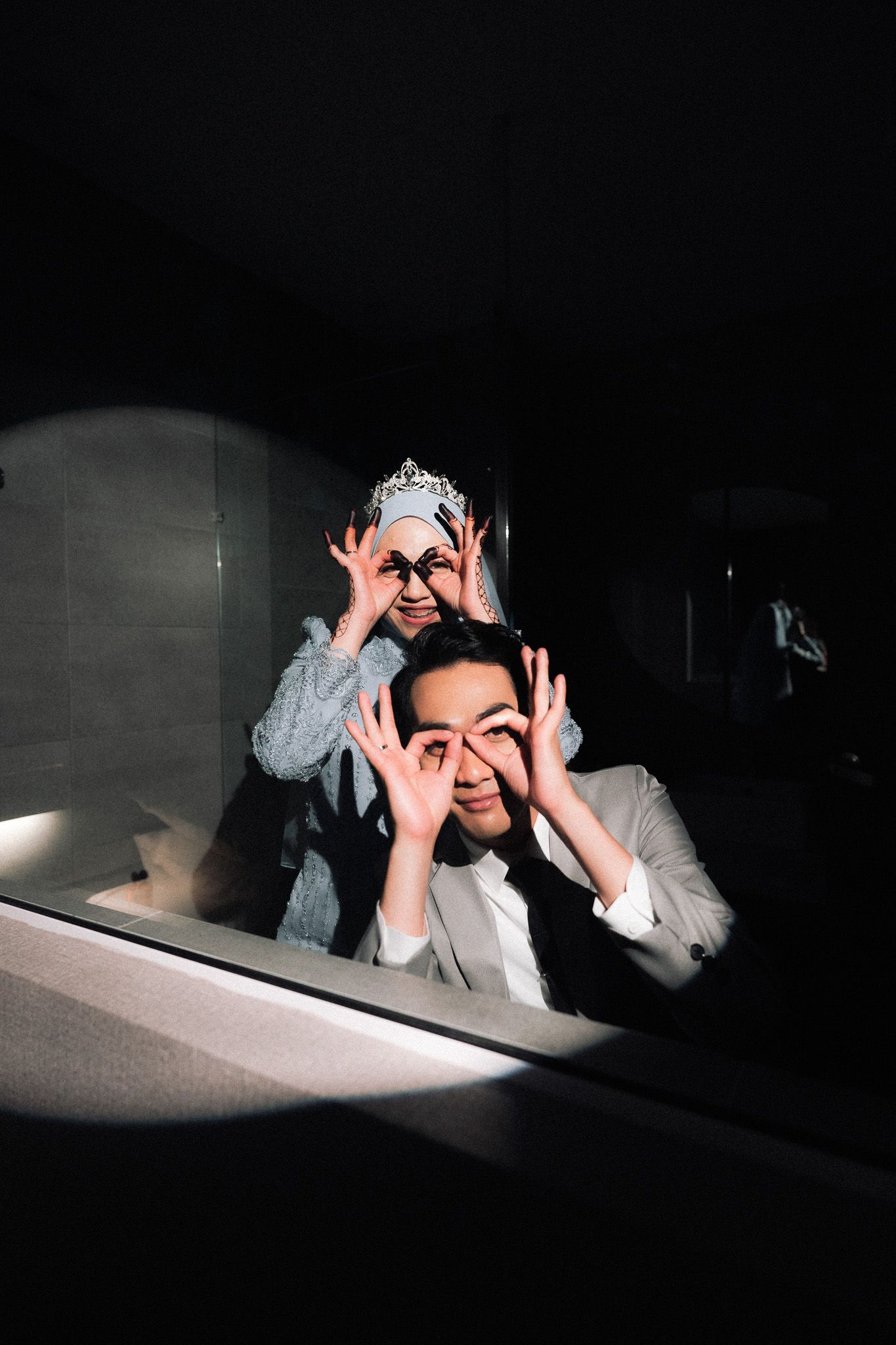 Couple making fun OK sign glasses gesture with fingers, woman wearing hijab and crown, man in suit, dark background.