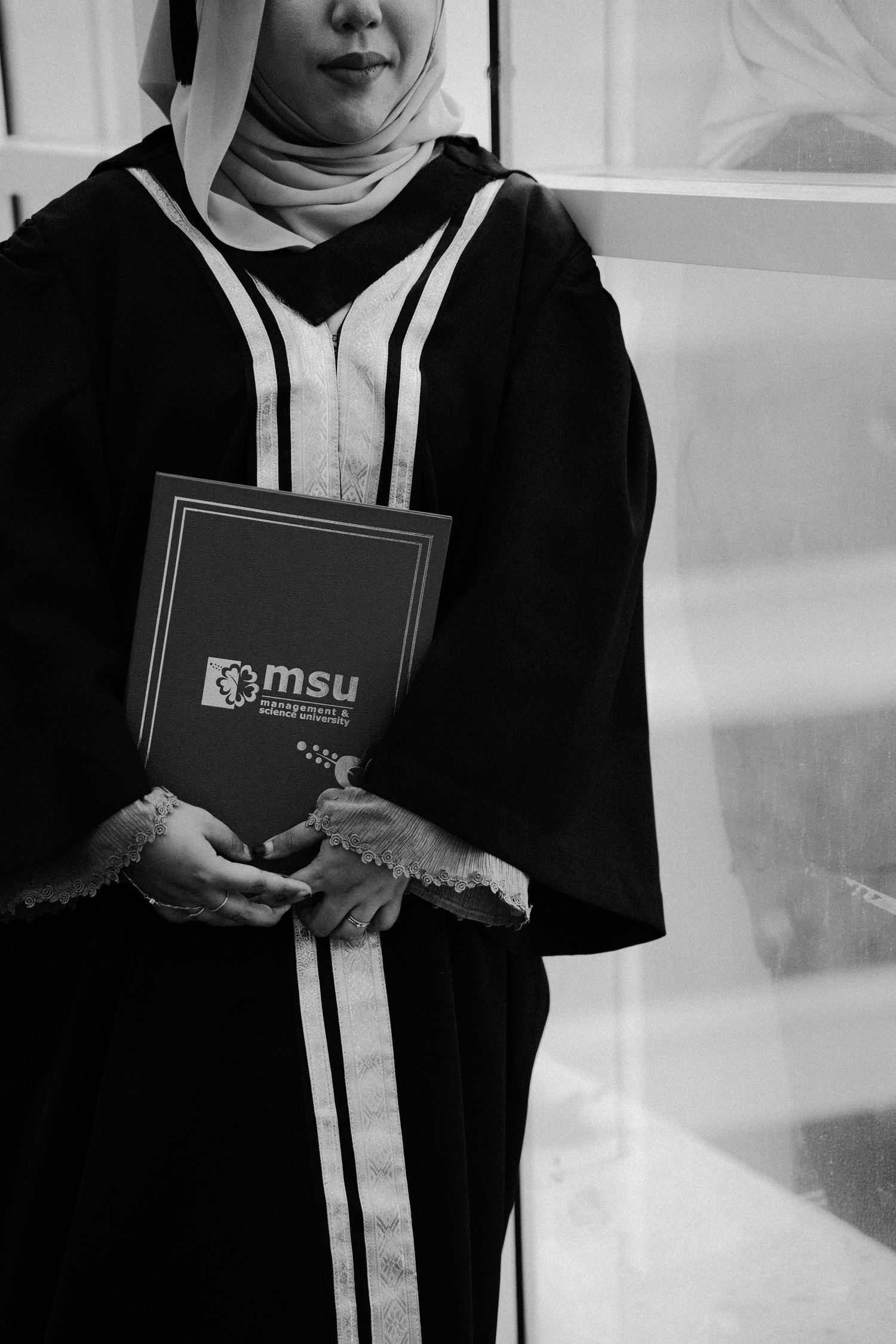 A graduate in black academic regalia holds an MSU handbook while wearing a white hijab in this black and white photo.