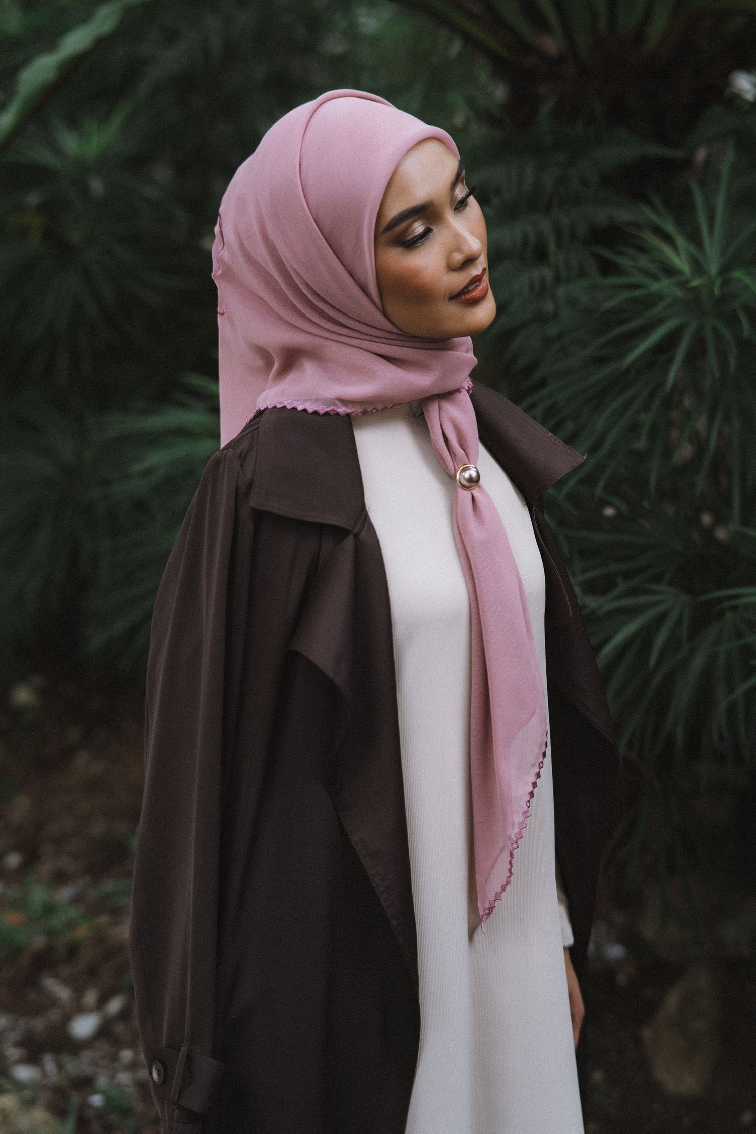 Elegant styling of pink hijab with brown coat and white dress against lush greenery.