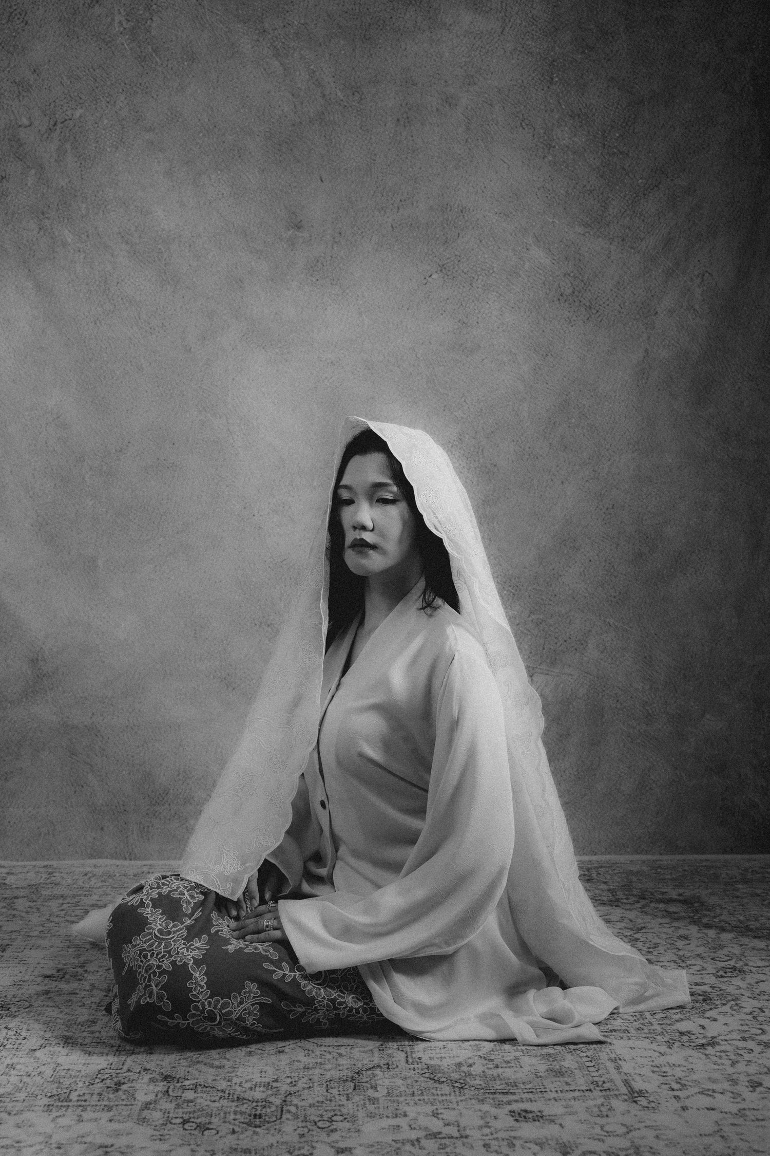 A monochrome studio portrait of a person in flowing robes and veil seated against a textured background.