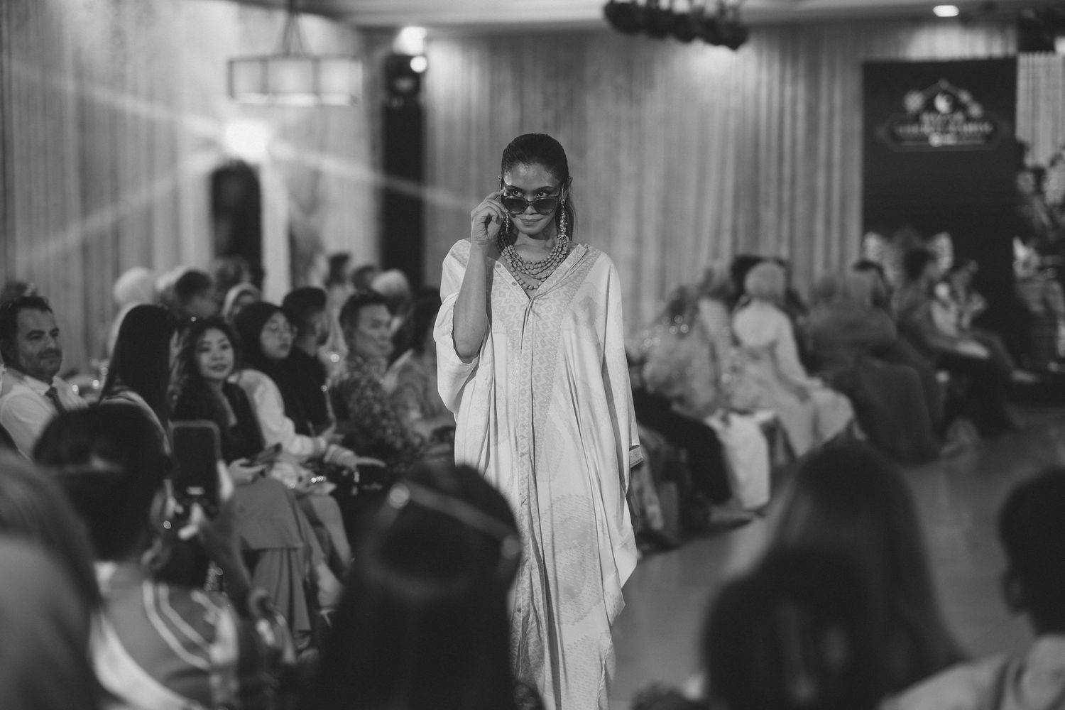 Black and white photo of model in flowing white dress on fashion runway.