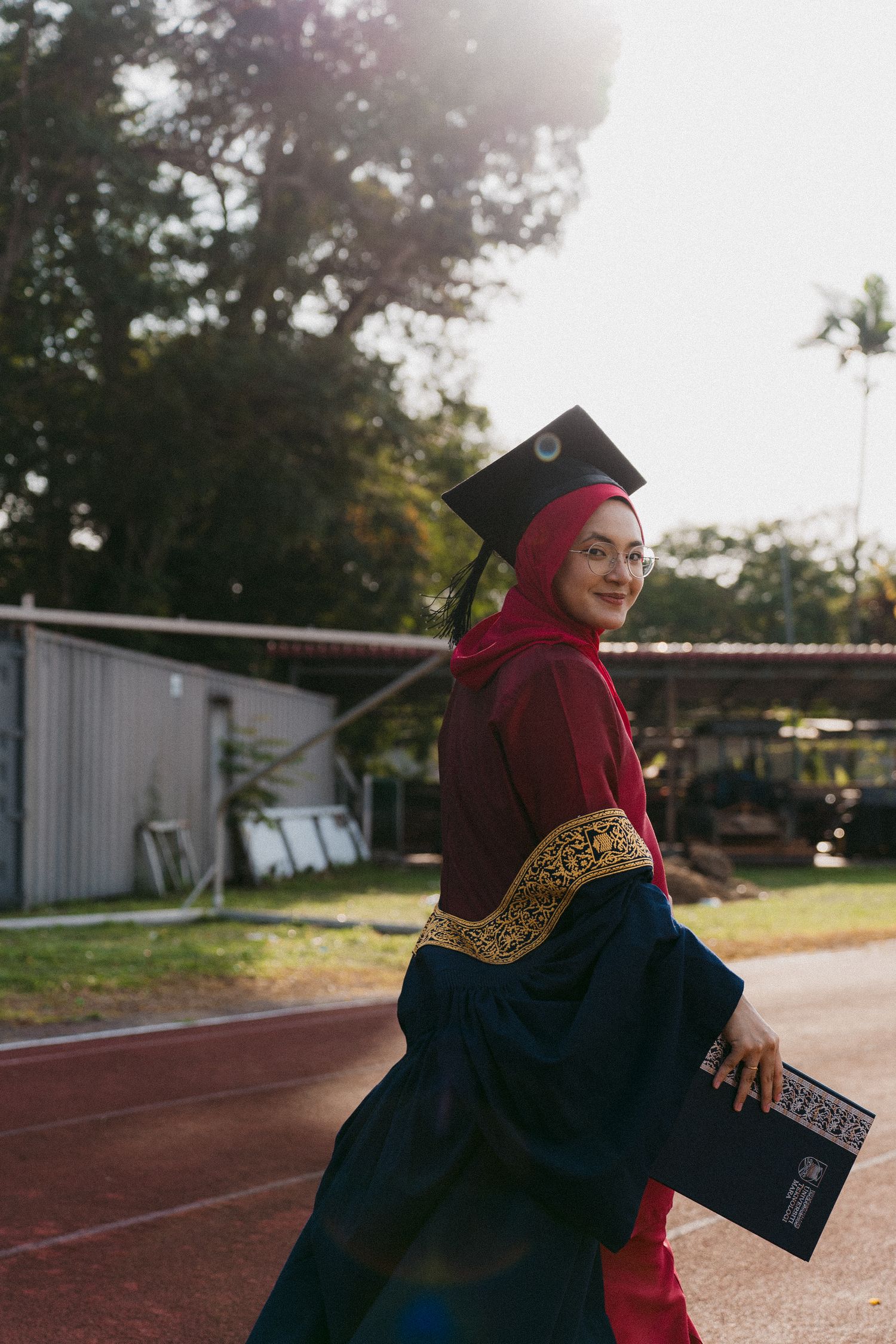 Graduate in traditional academic regalia and red hijab poses with diploma on outdoor running track.
